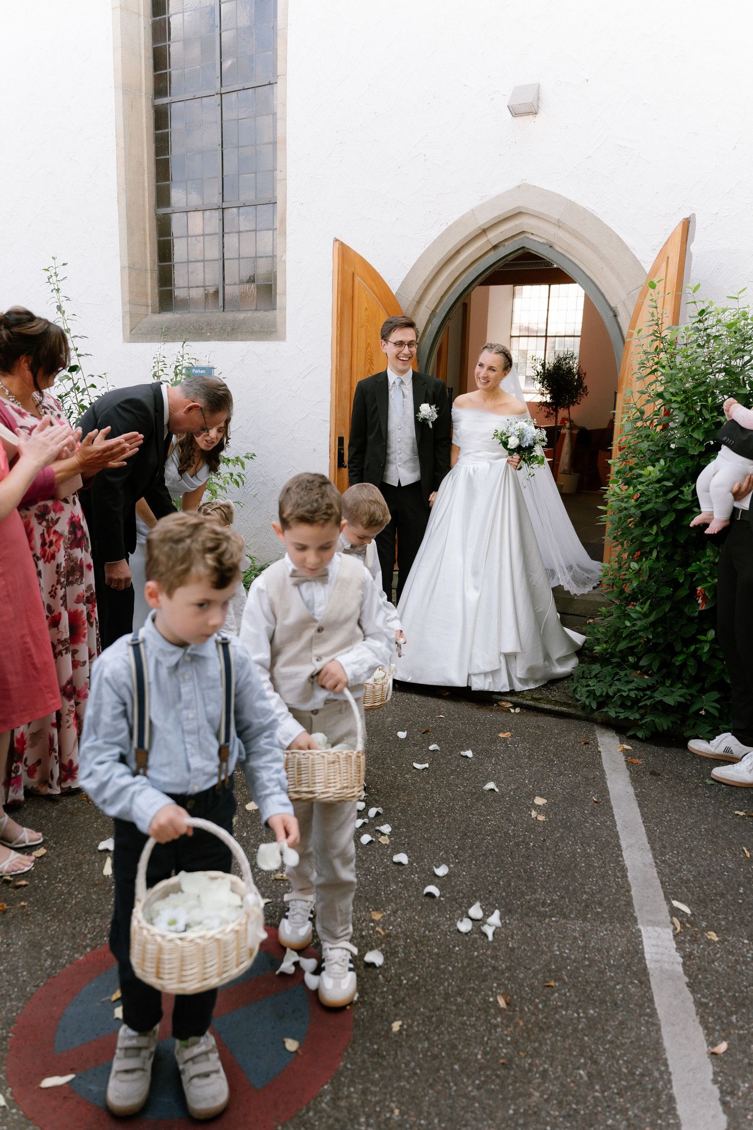 Bride and groom smile in a church doorway as flower boys toss petals from wicker baskets while wedding guests applaud.