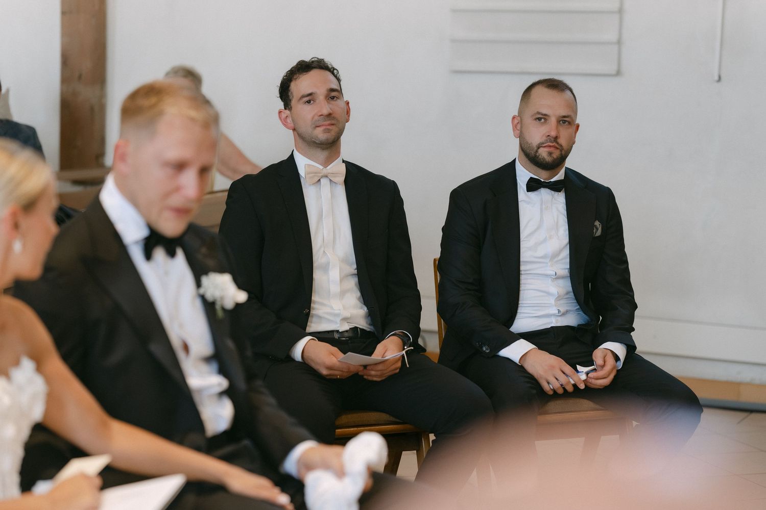 Three men in dark suits and bow ties sit attentively in a row during a wedding ceremony, with a woman in white at the left edge.