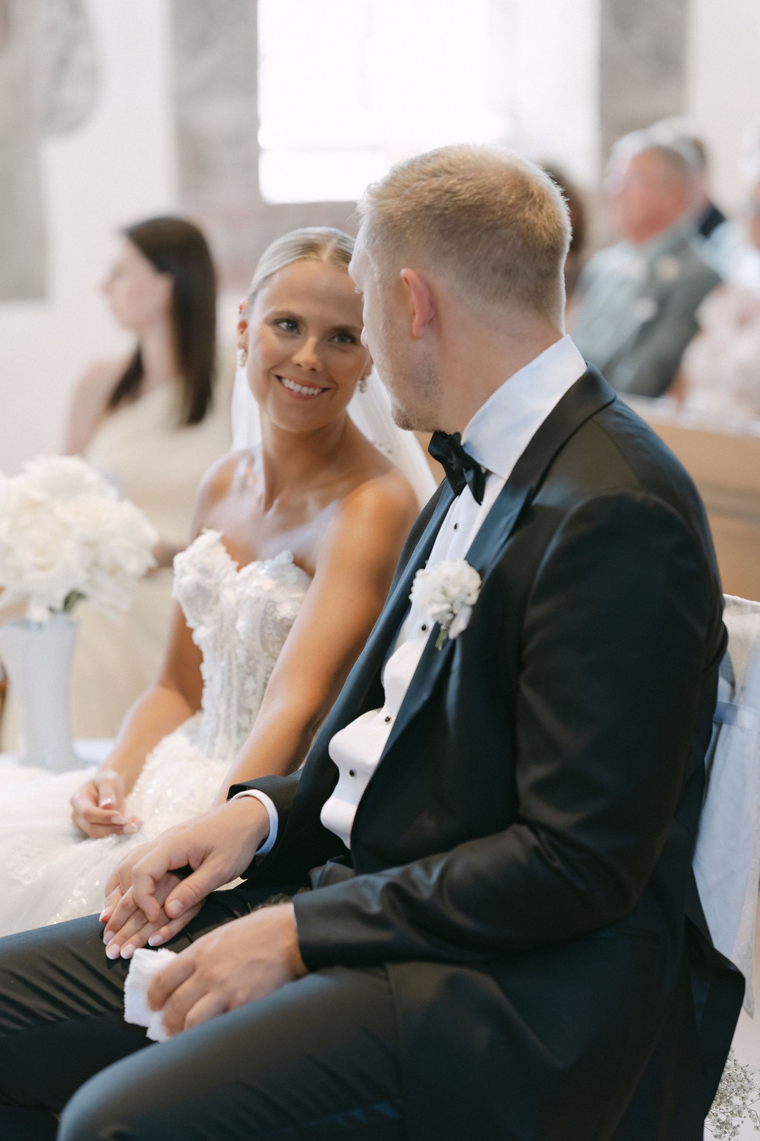 Smiling bride in a lace wedding gown gazes at her groom in a black tuxedo with bow tie as they hold hands during a church ceremony.