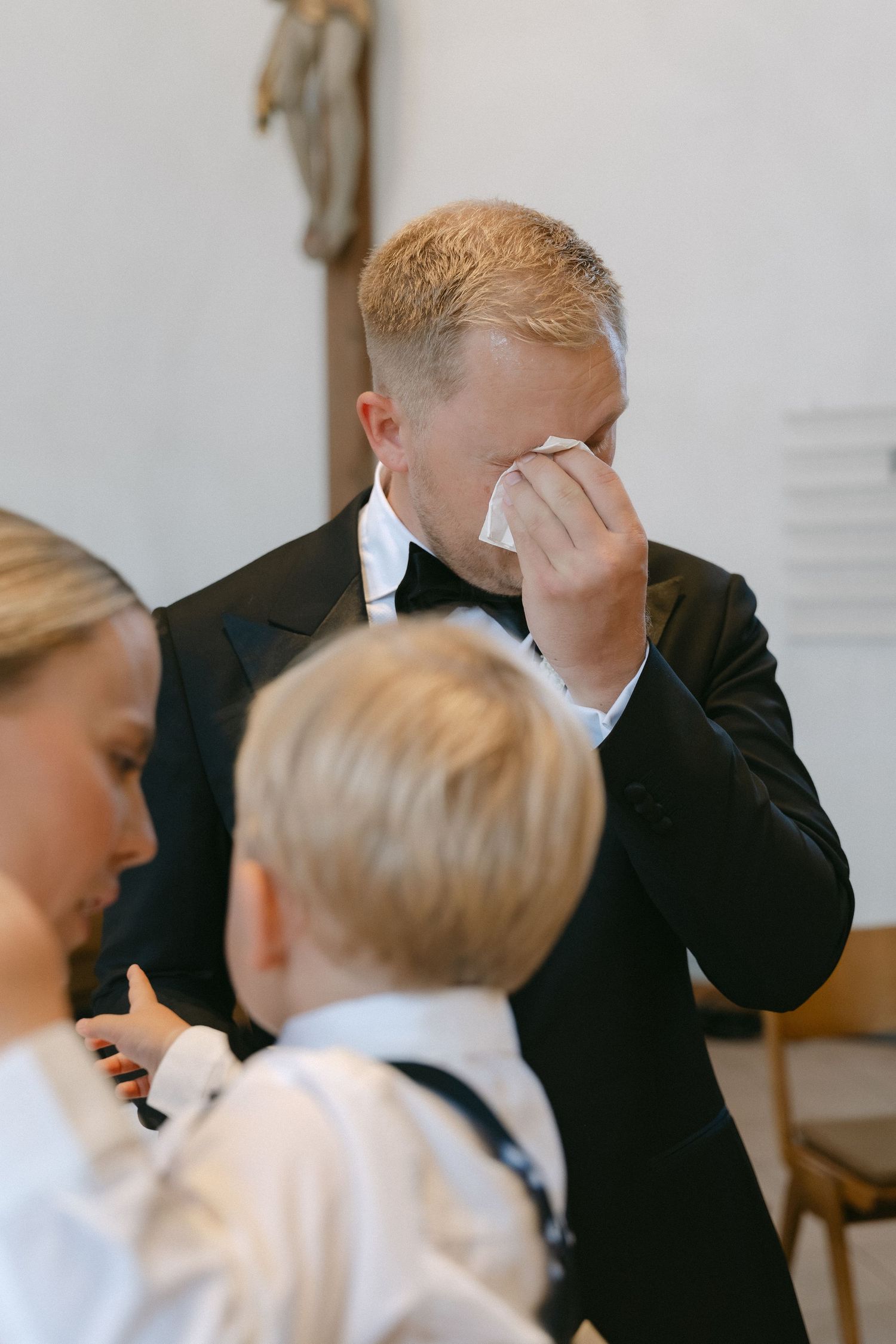 A groom in a black tuxedo and bow tie wipes away tears with a tissue during a wedding ceremony while a young boy and woman stand nearby.