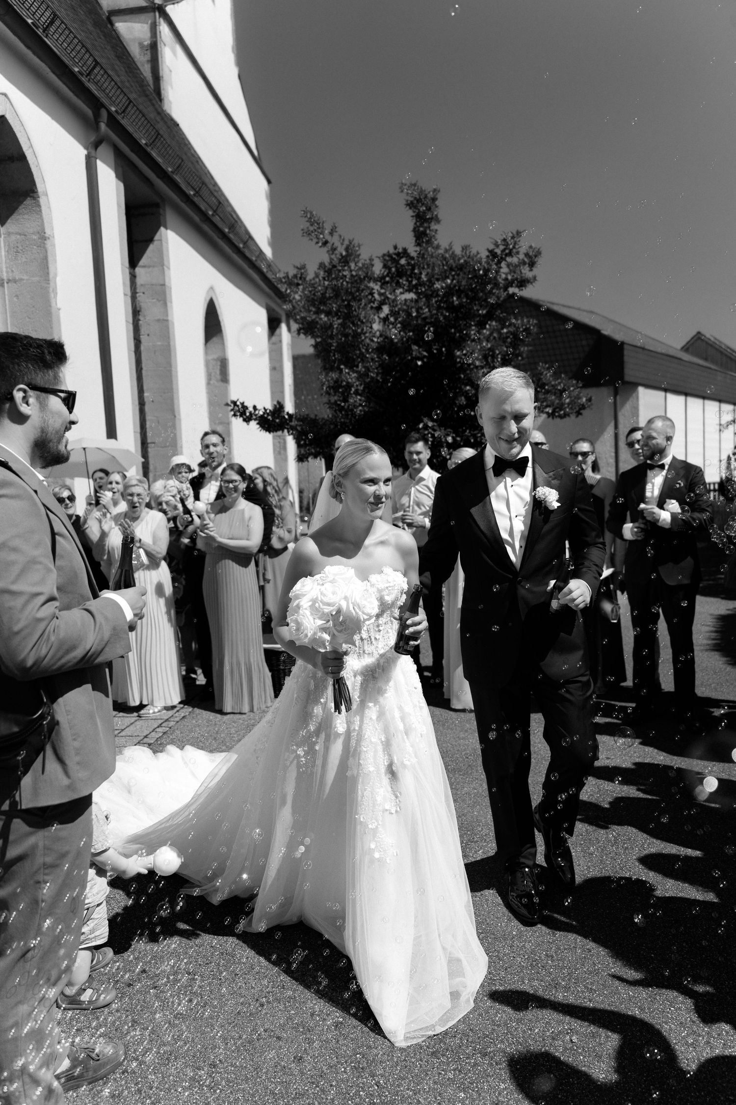 Bride in a lace gown with bouquet and groom in a bow tie walk past cheering wedding guests outside a church in black and white.