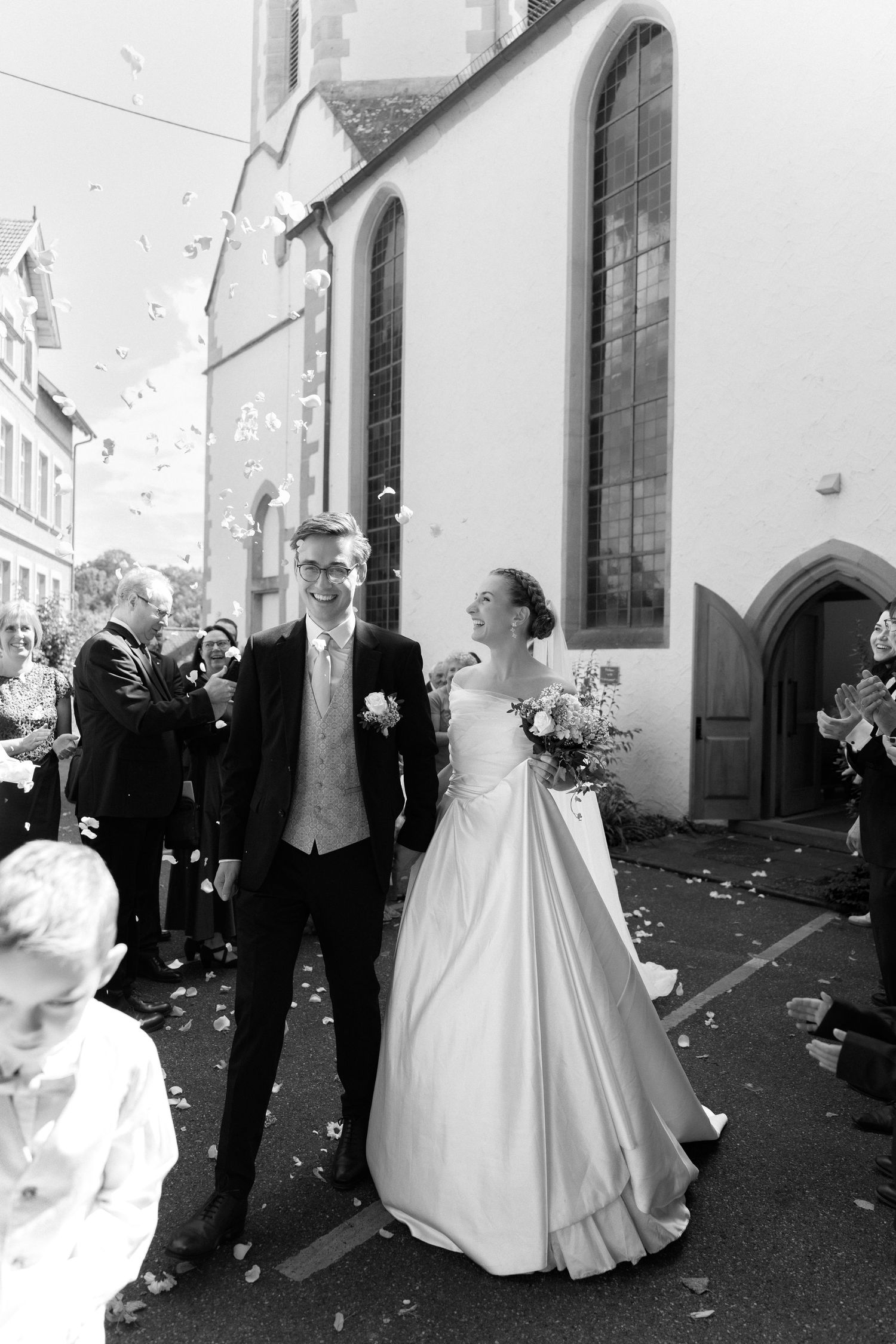 Smiling bride and groom walk outside a church as guests toss confetti in a joyful black and white wedding photo.