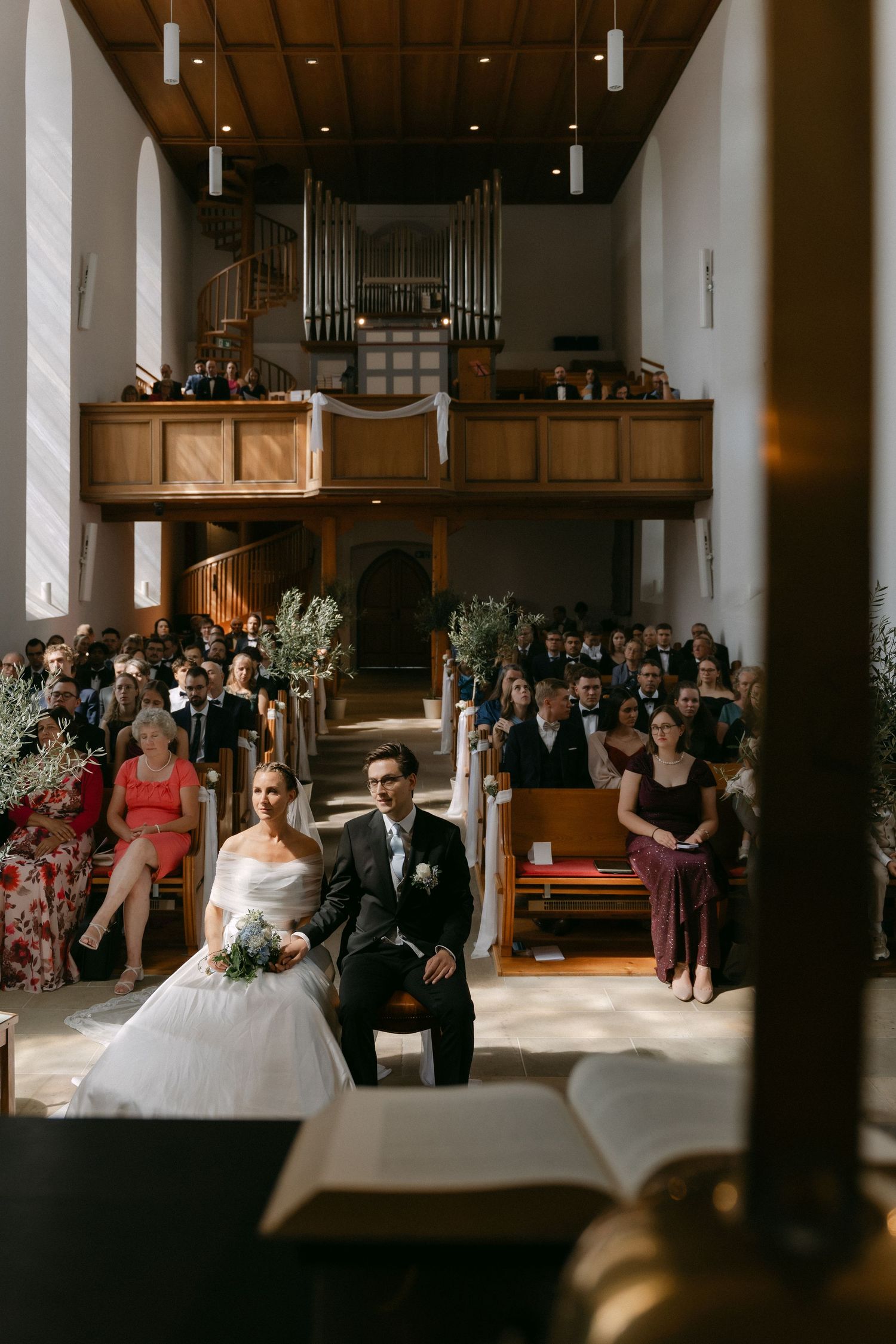 Bride in a white gown and groom in a dark suit sit facing the altar during a church wedding ceremony as guests fill the wooden pews.