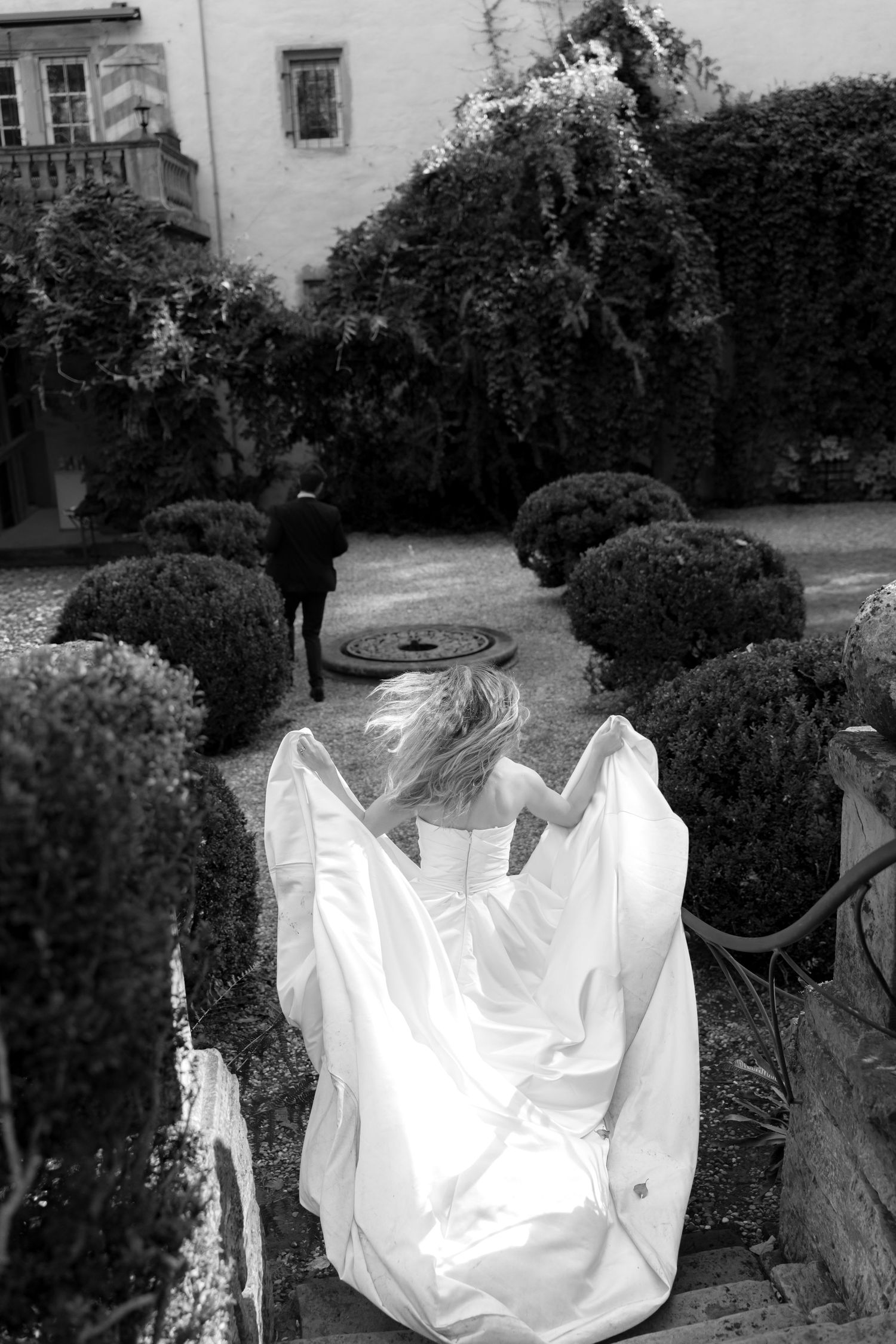 Bride in a flowing white wedding gown walks down garden steps toward a groom in a dark suit, in black and white.