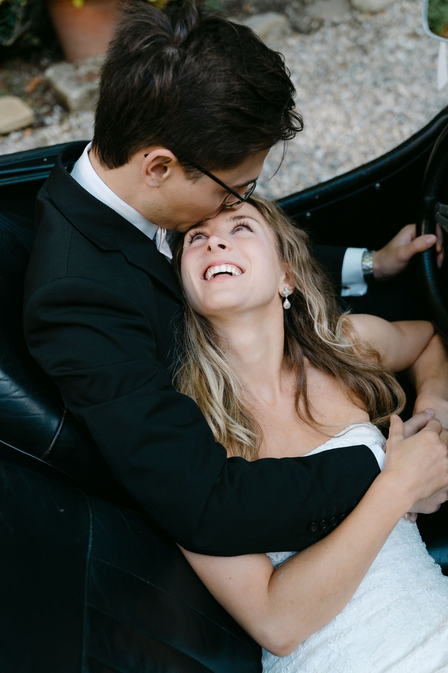 Groom in a black suit and glasses kisses the forehead of a laughing bride in a white lace wedding dress while seated in a vintage car.