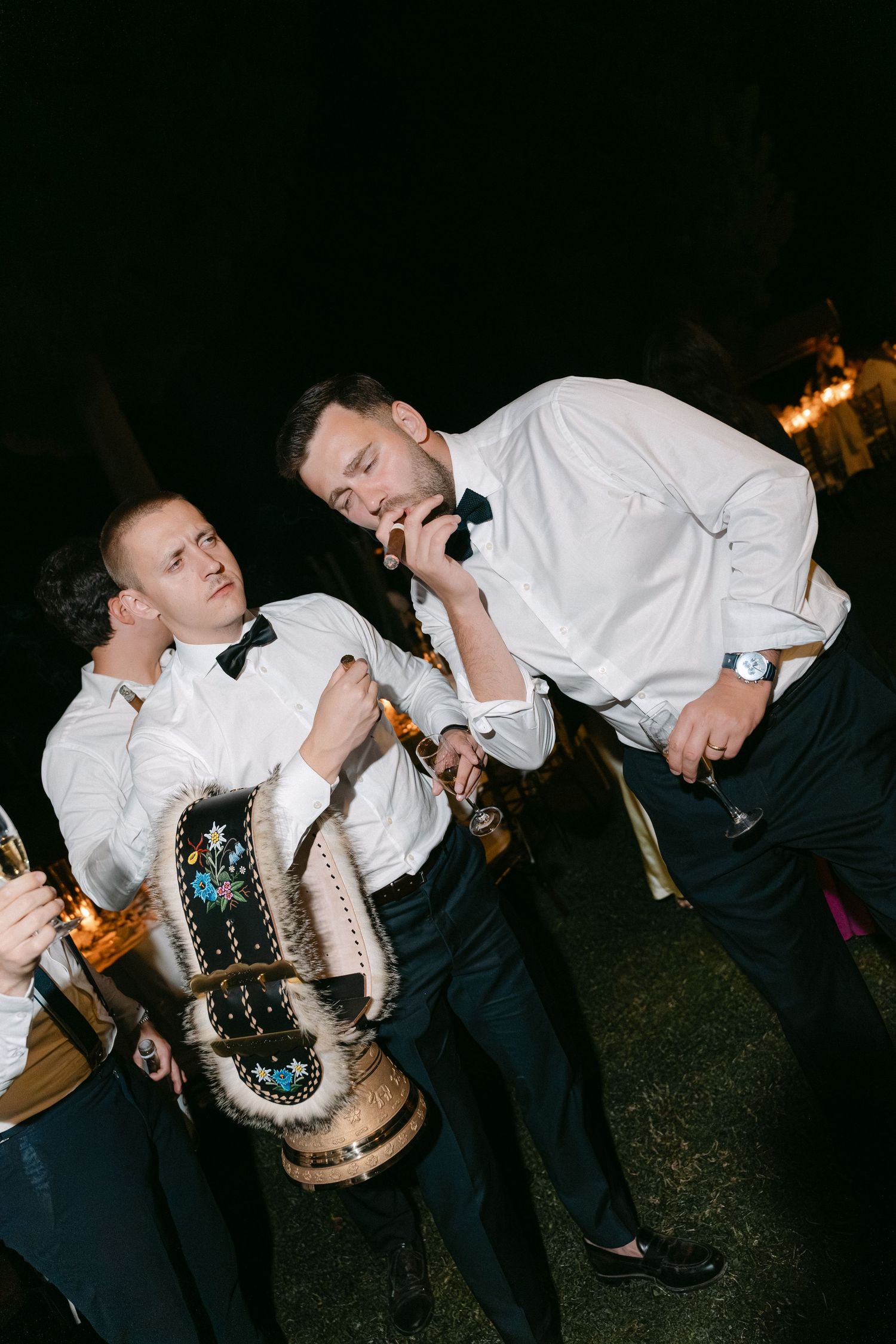Two men in white shirts and bow ties celebrate at a nighttime event, one smoking a cigar while holding drinks.