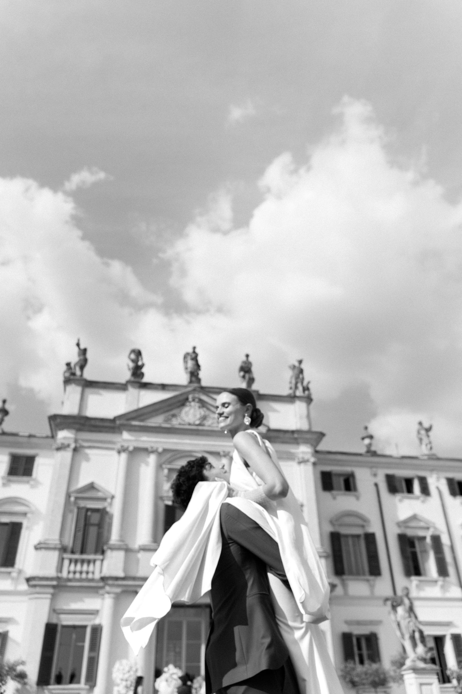 Black and white photo of statues atop white buildings against a cloudy sky in an architectural cityscape. Villa Mosconi Bertani