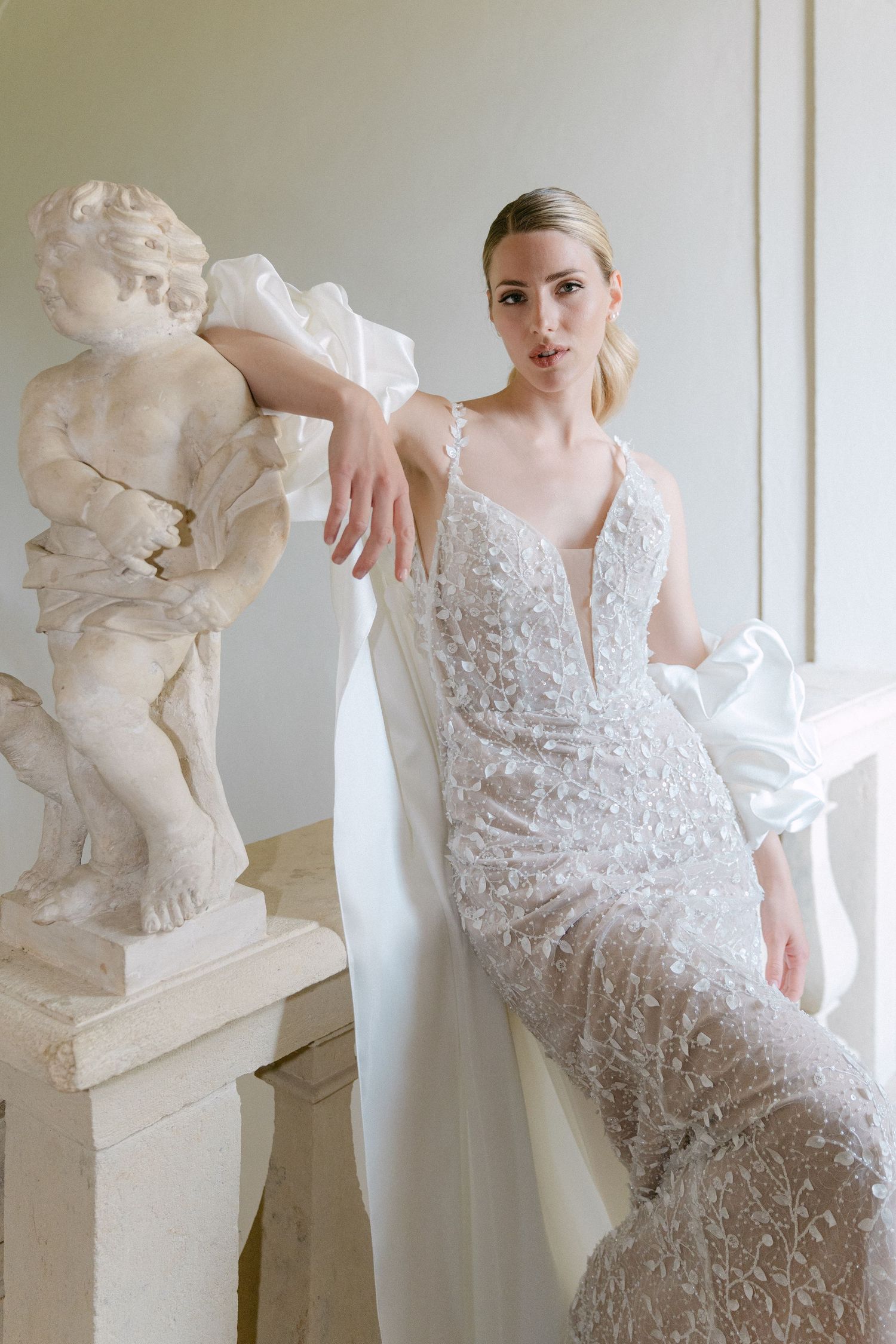 Elegant bridal gown with delicate floral embroidery and plunging neckline posed next to classical cherub statue. Wedding Verona