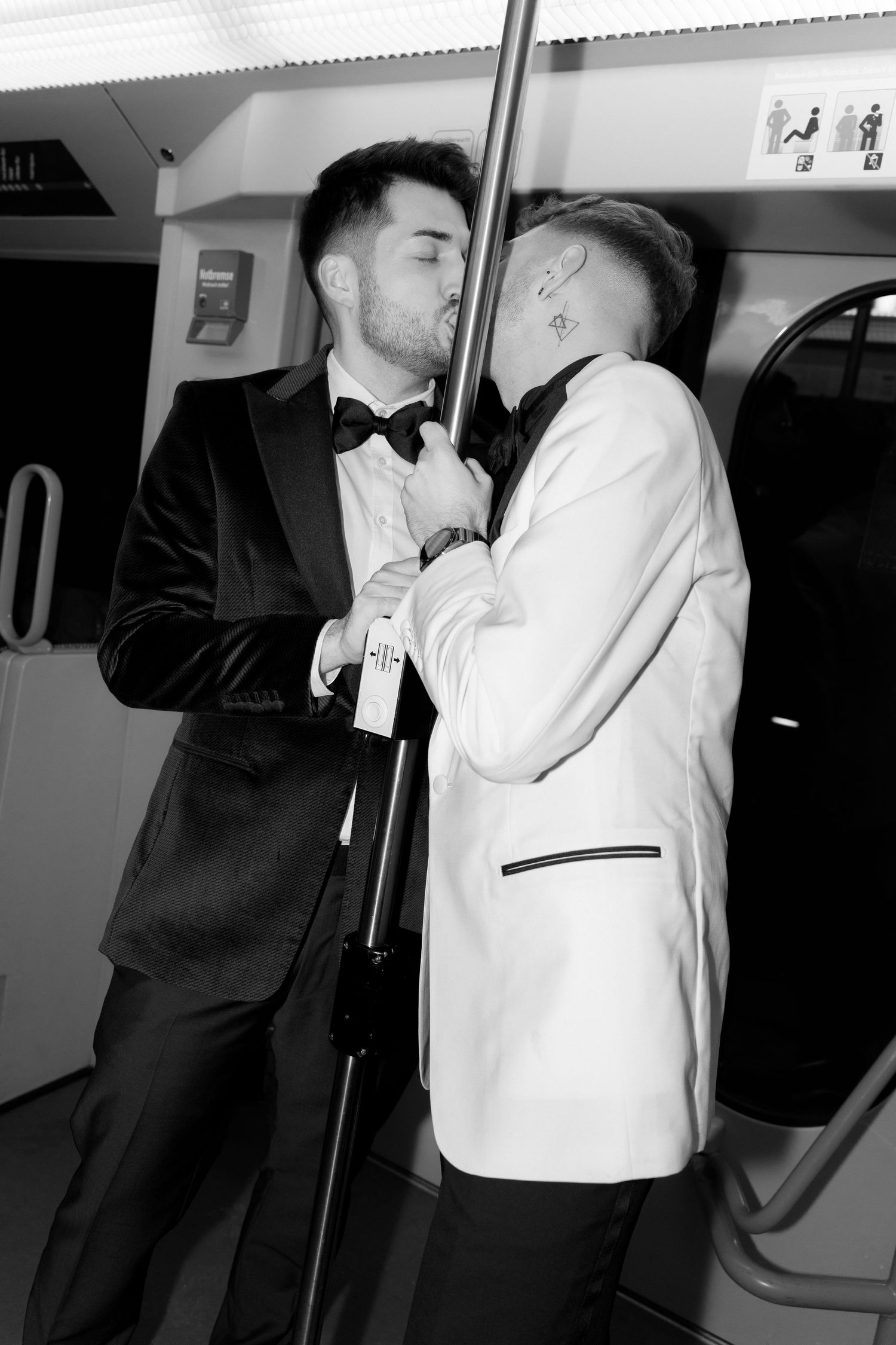 Two people in formal attire, one in black tuxedo and one in white jacket, standing together near a pole in black and white photo.