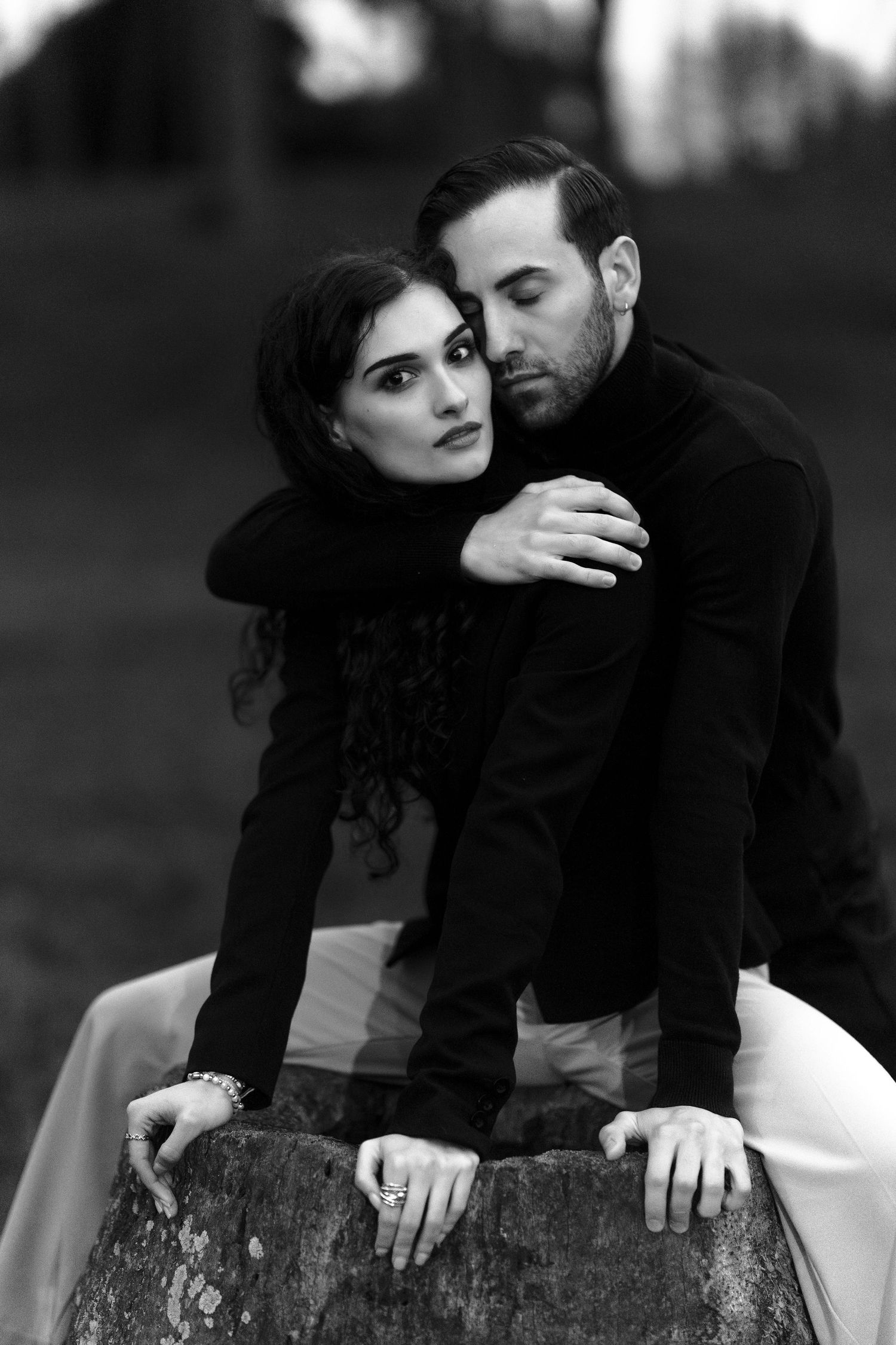 A romantic black and white portrait of a couple in dark clothing embracing on a rustic wooden surface outdoors.