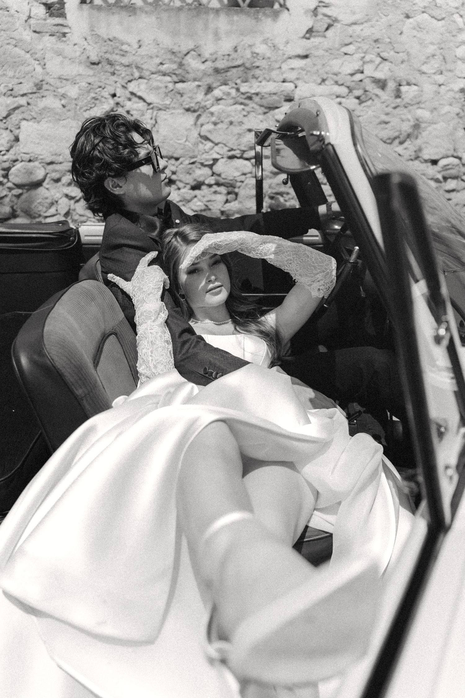 A vintage black and white photograph showing two people relaxing in a convertible car on a sunny day.