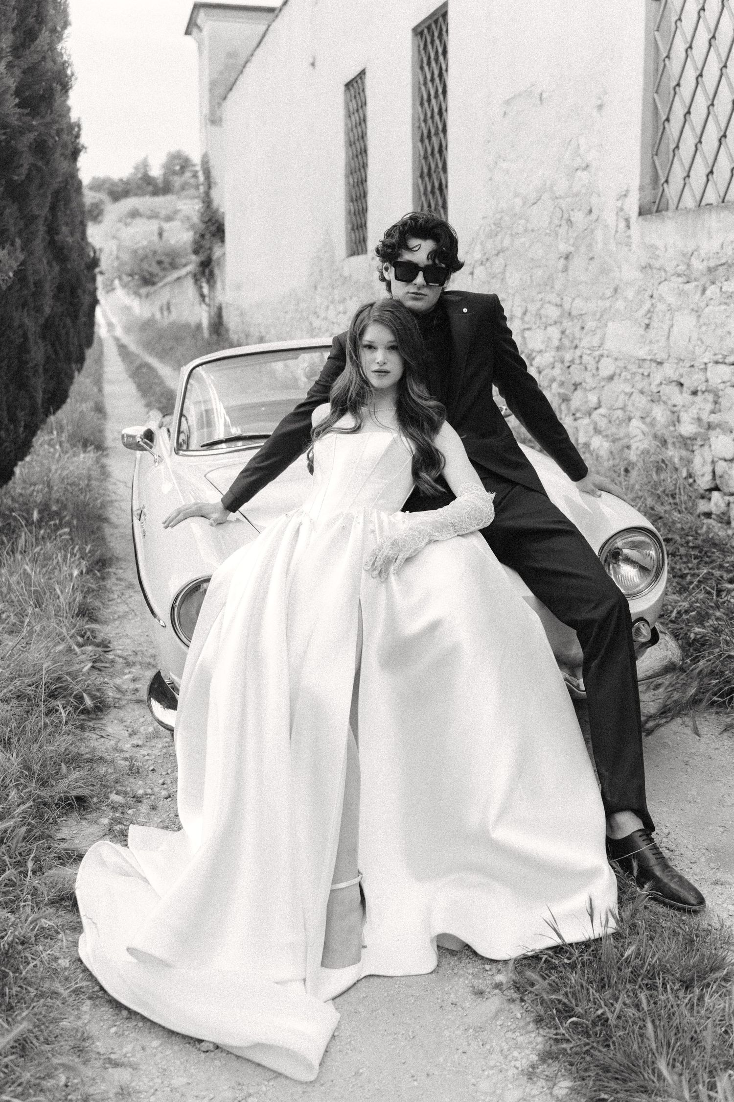 A black and white vintage-style wedding photo with a couple posing on a classic car against a white building.