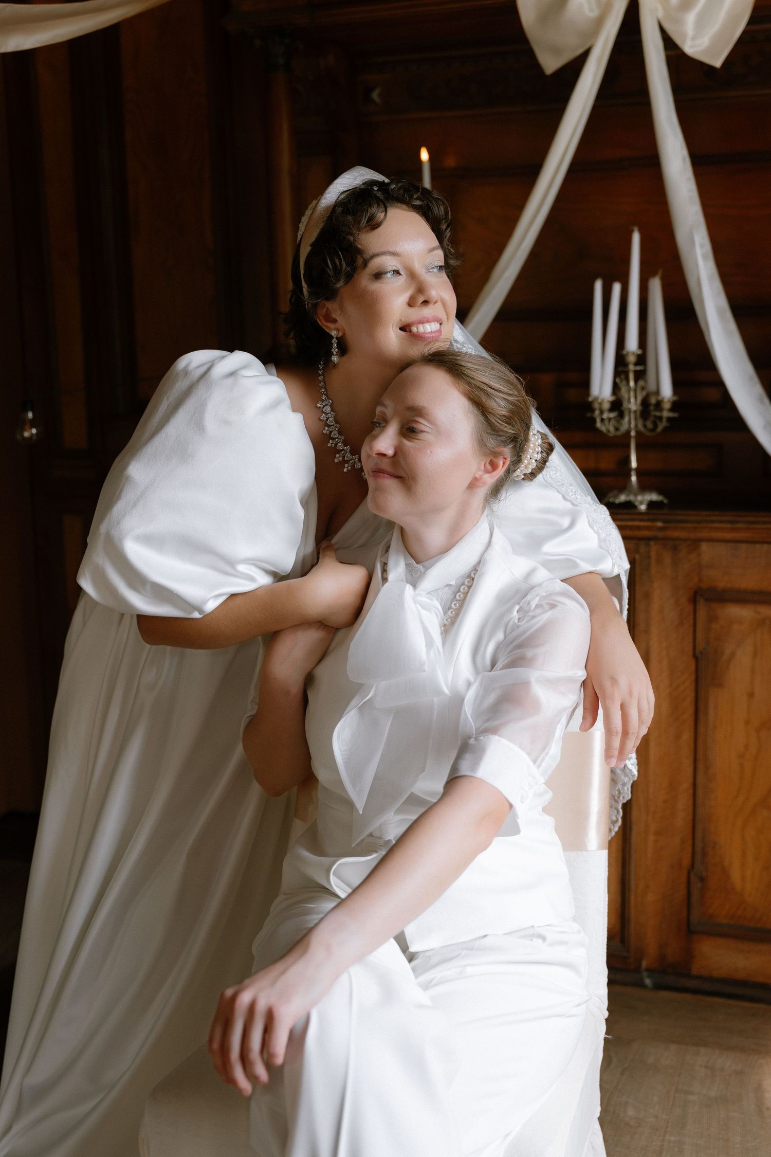 Two brides in white wedding gowns embrace warmly in an elegant wood-paneled room with candles.