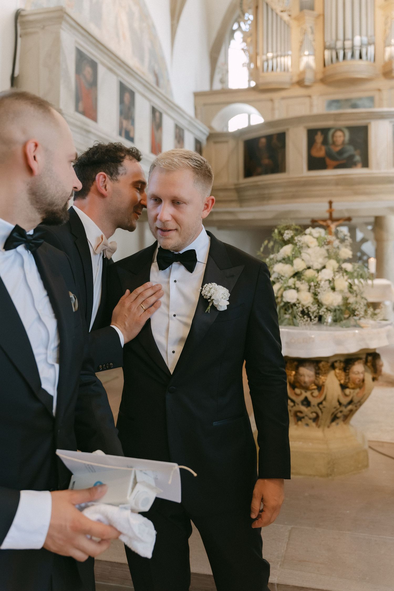 Groom in black tuxedo with bow tie and white boutonniere listens as a groomsman whispers in his ear inside a church with floral decor.