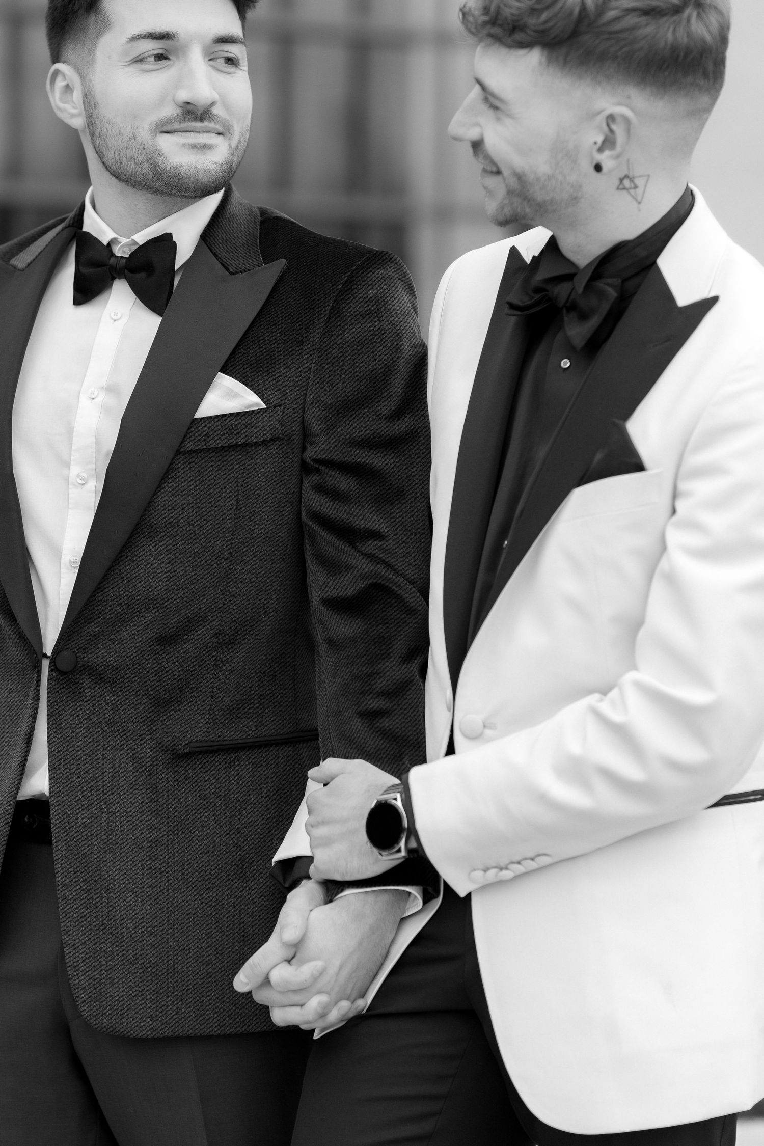 A black and white photo showing two people in formal attire, one in black tuxedo and one in white jacket, holding hands.