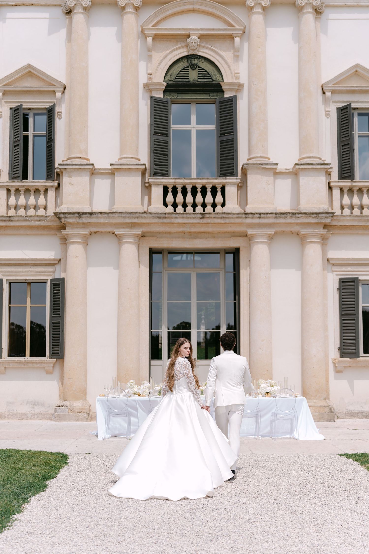 Elegant wedding couple stands in front of a grand Italian villa with classic architecture and outdoor table settings. Villa Mosconi Bertani