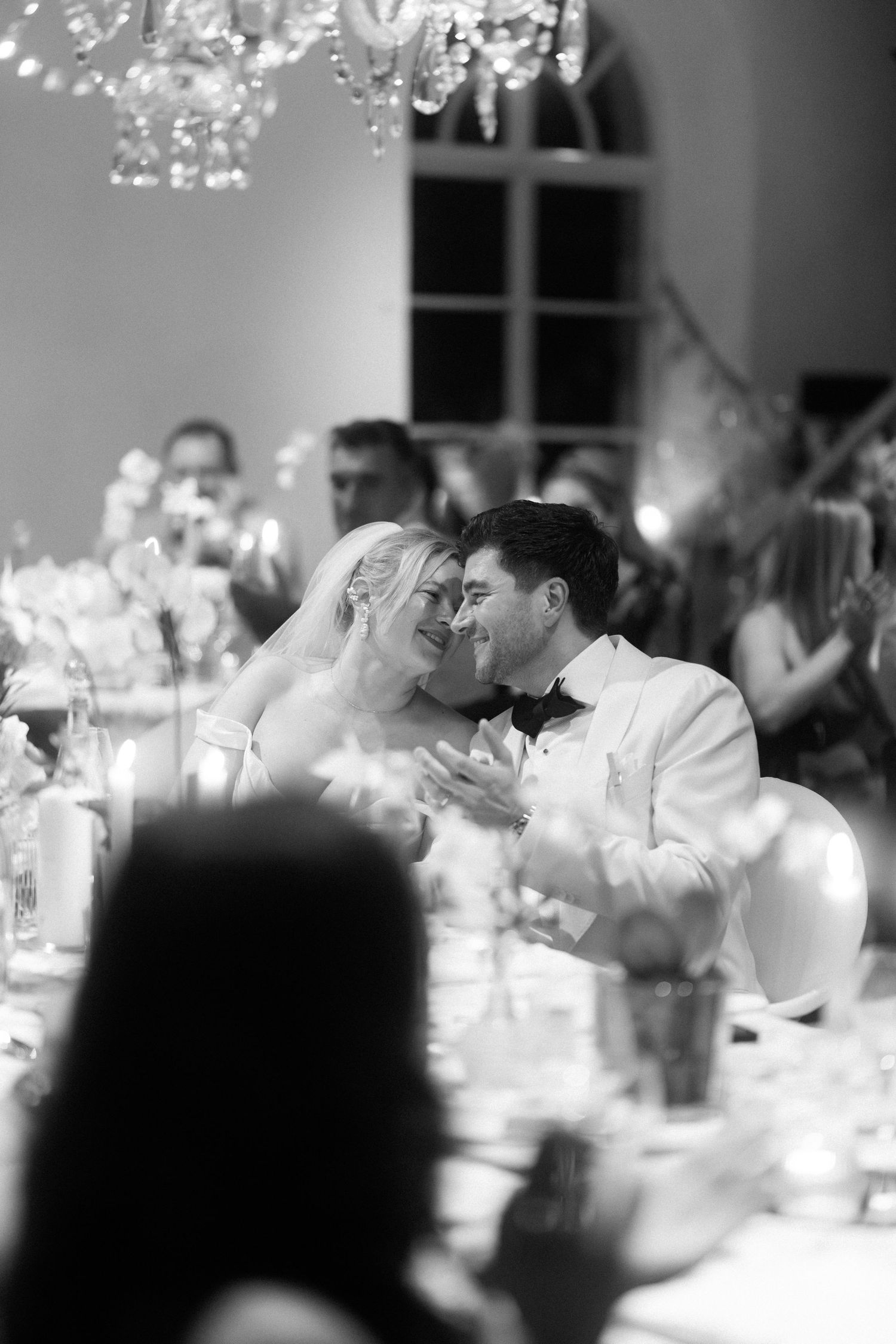 A tender black and white wedding reception moment captured at an elegant dinner table under crystal chandeliers.