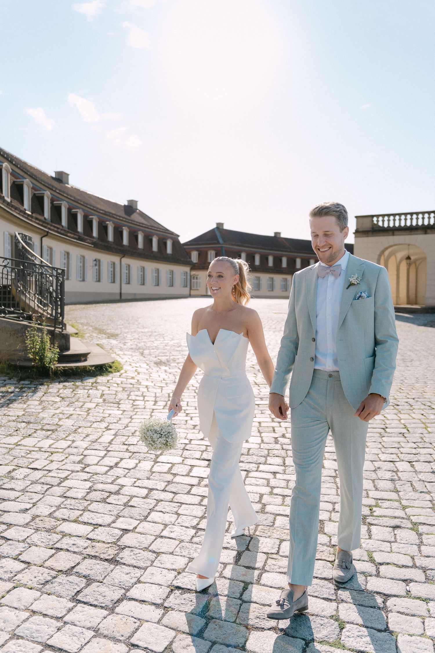 A couple in wedding attire walks hand in hand on a cobblestone courtyard of a historic European building on a sunny day. Schloss Solitude