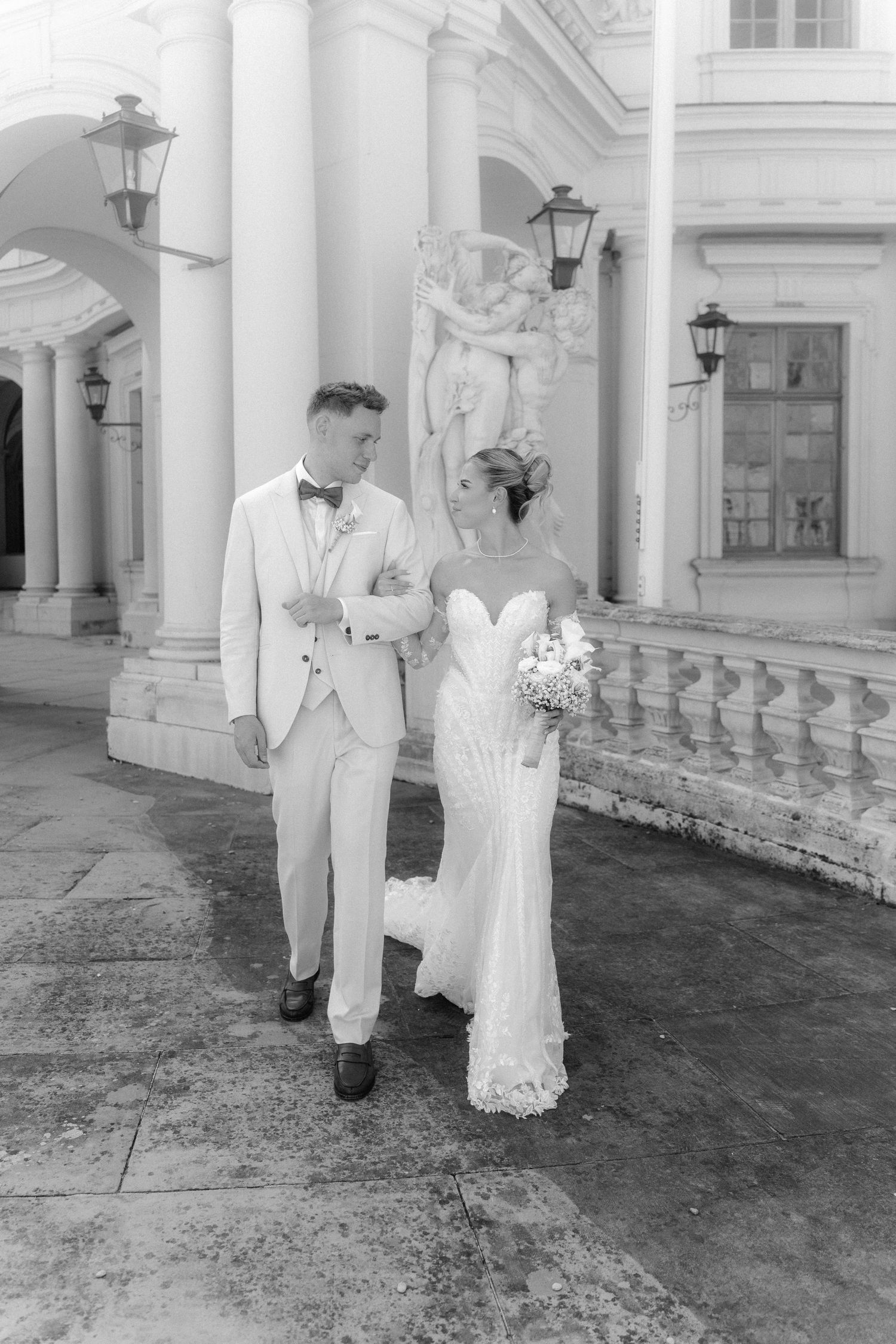 A couple in elegant wedding attire pose together on stone steps against a classical architecture backdrop with columns. Schloss Ludwigsburg