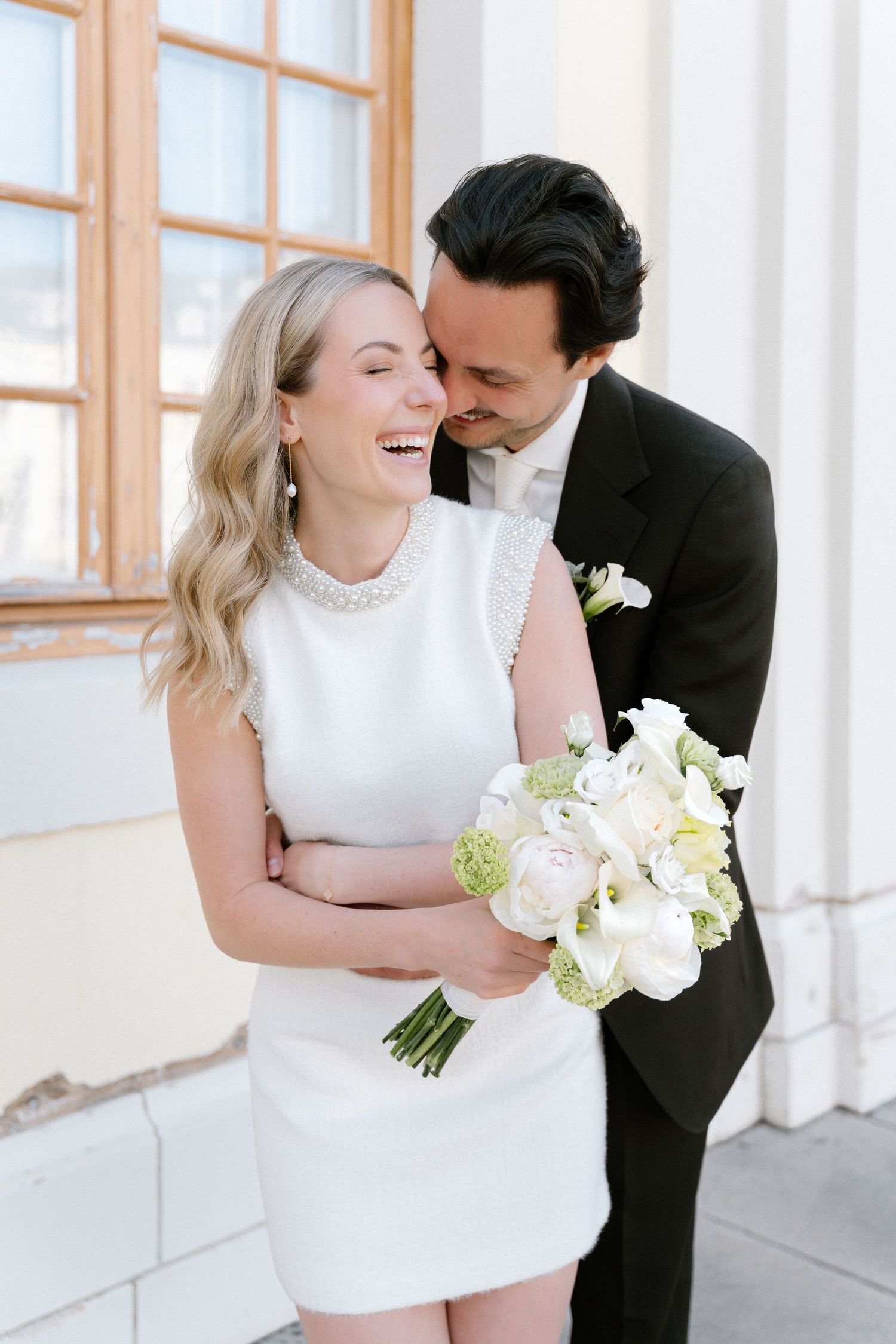 Joyful bride in white mini dress holding bouquet embraced by groom in black suit at elegant outdoor venue.
