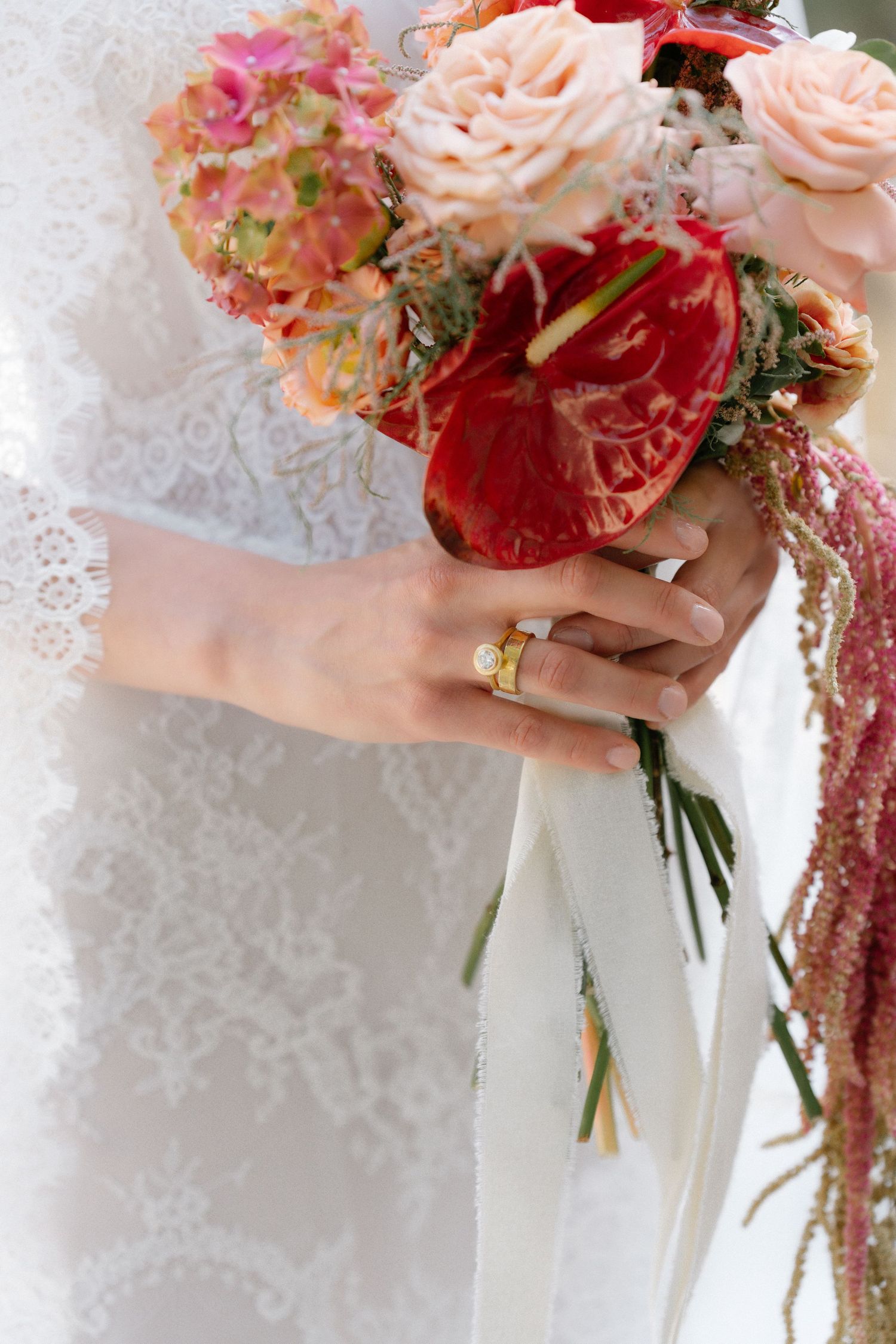 Bride in white lace dress holding a colorful wedding bouquet with red anthuriums and pink roses, wearing a gold ring.