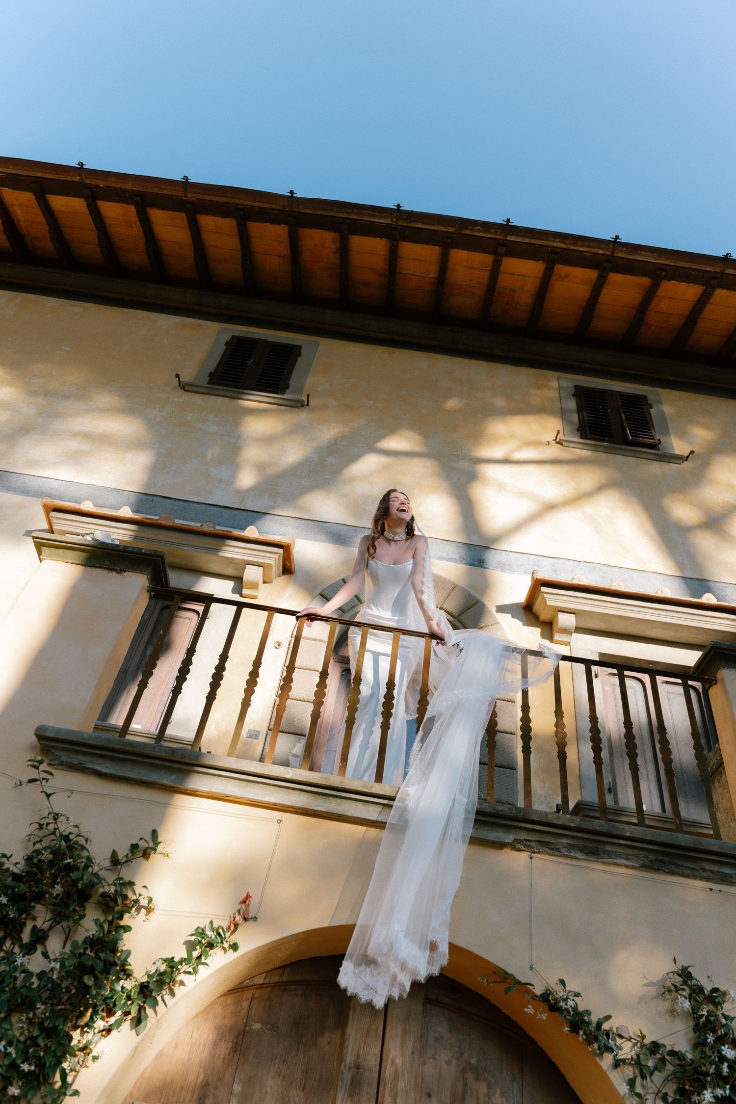 Bride in white lace gown standing on sunlit balcony of rustic Italian villa, veil flowing over railing.
