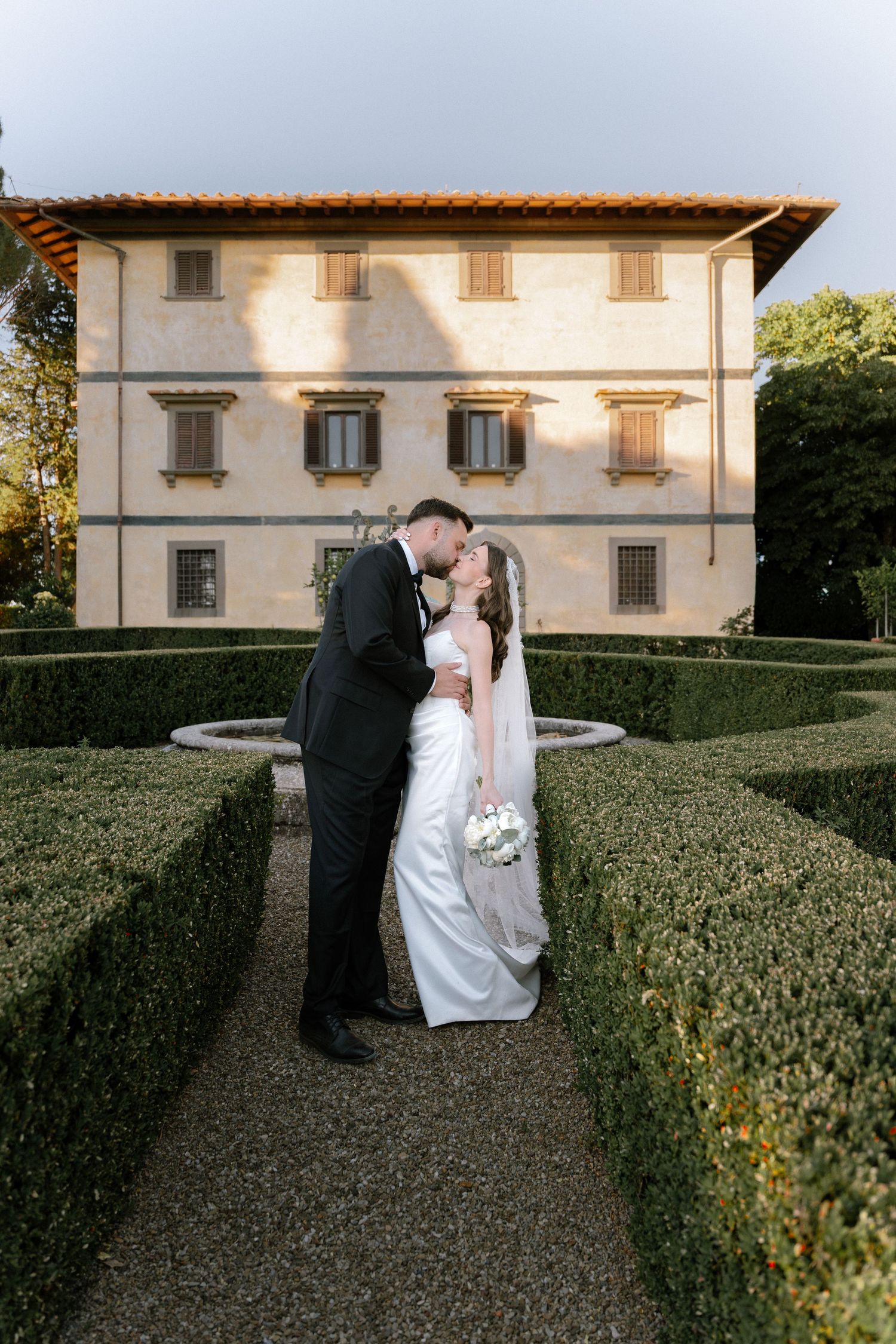 Bride and groom kissing in a formal garden maze with an elegant Italian villa in the background.