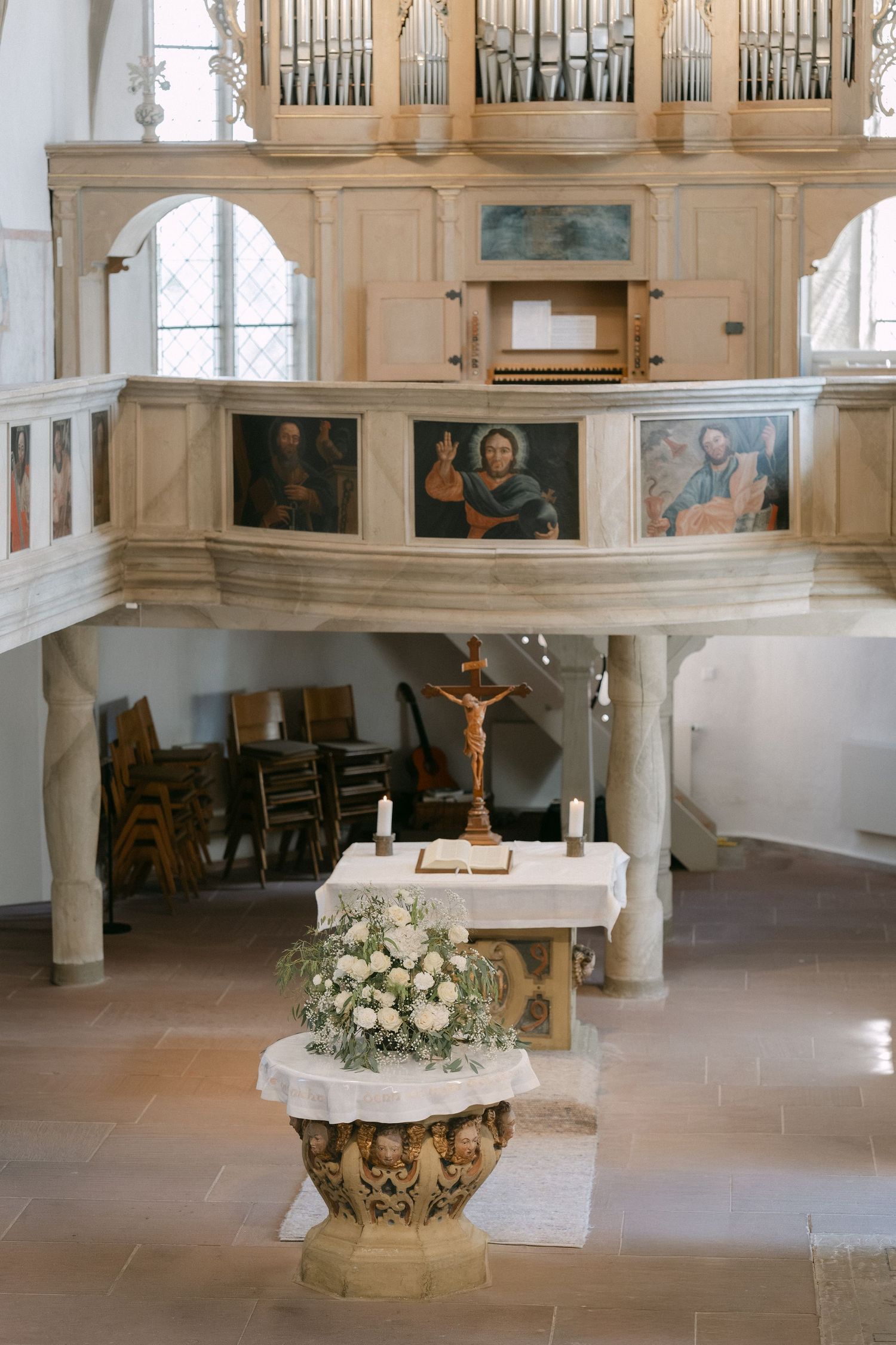 Church interior with a floral-decorated baptismal font, altar with crucifix and candles, and a painted organ gallery above.
