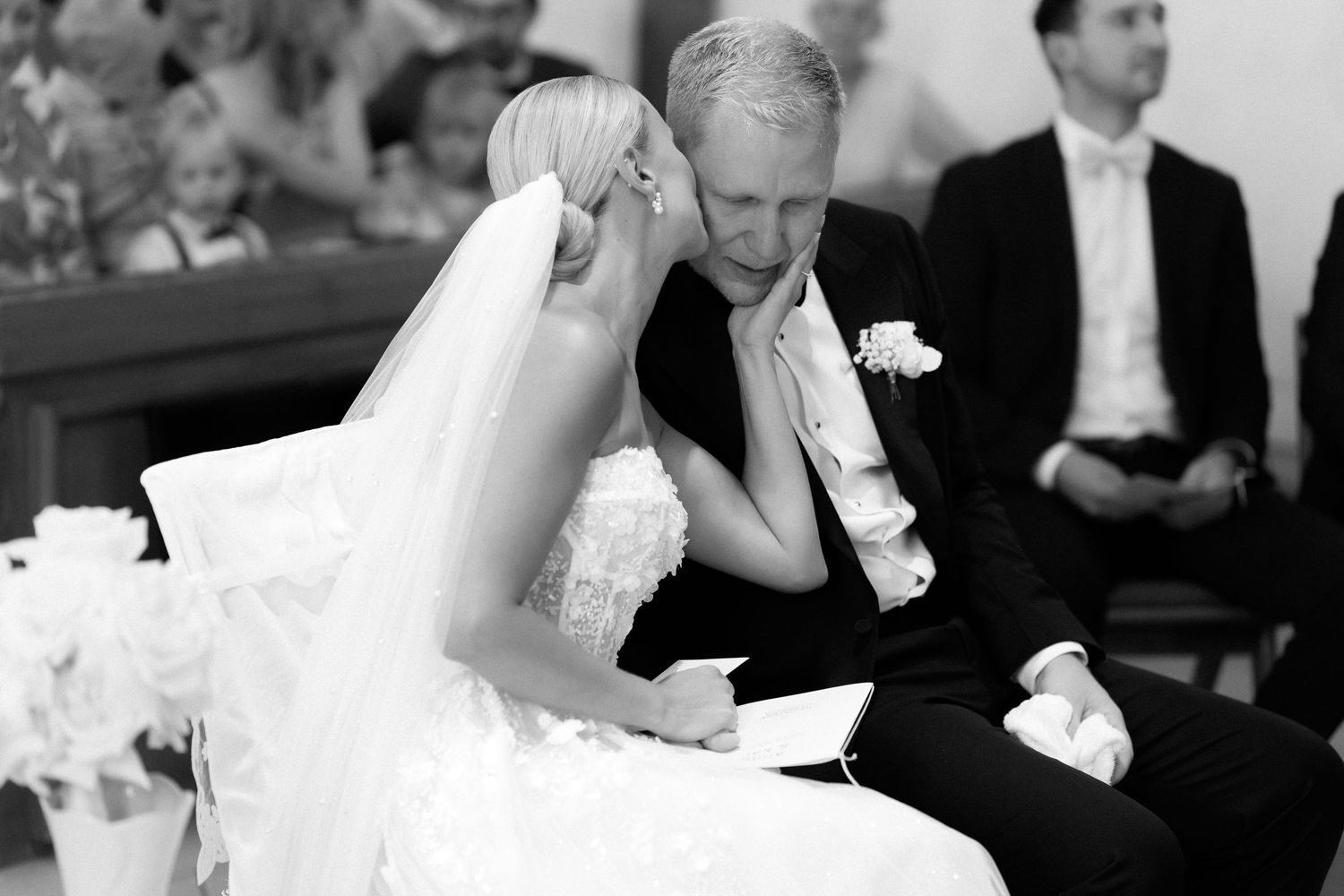 Black and white photo of a bride in a lace gown and veil whispering to an older man with a boutonniere during a wedding ceremony.