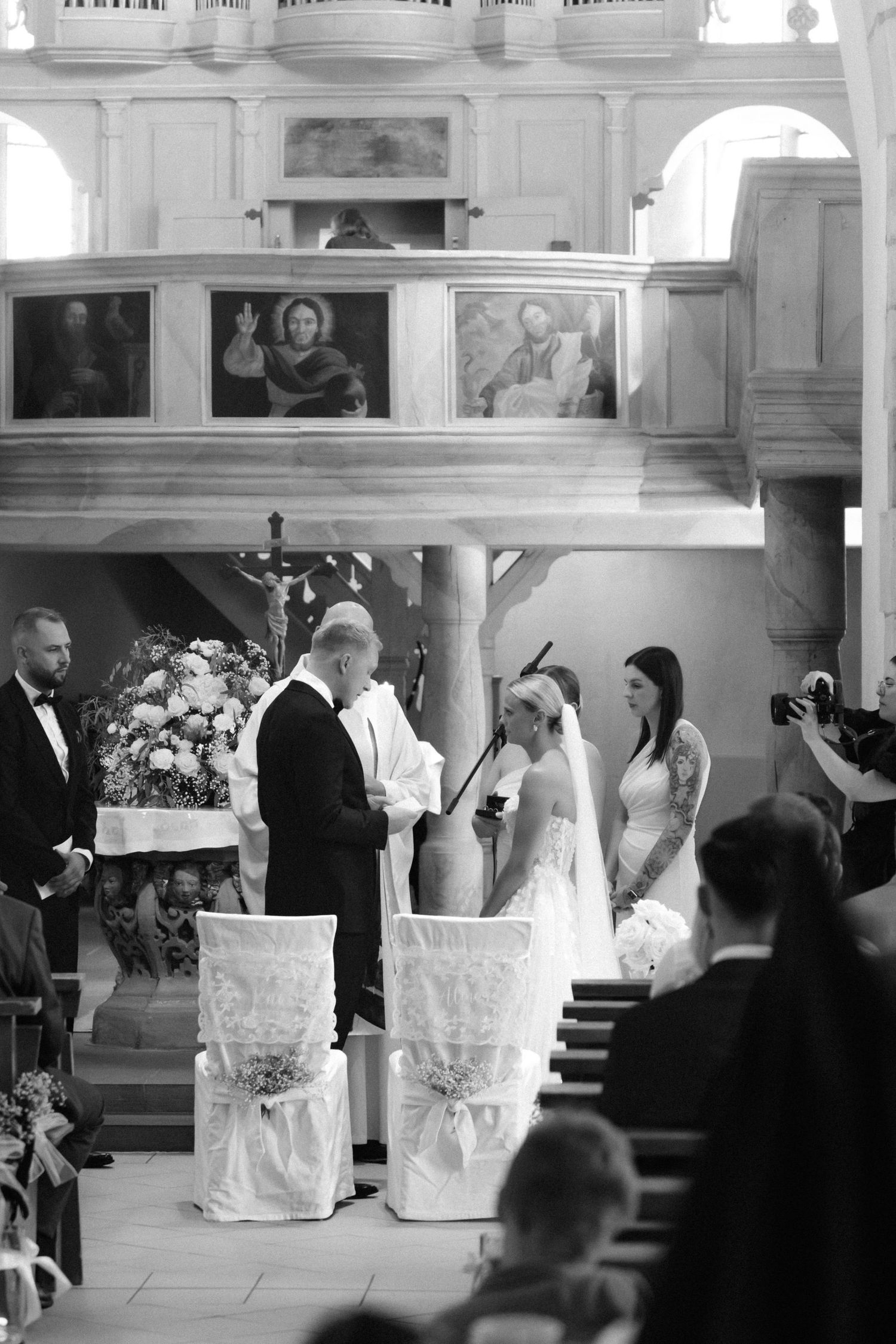 Black and white photo of a bride and groom exchanging vows before a priest in a church, with floral decor and guests looking on.