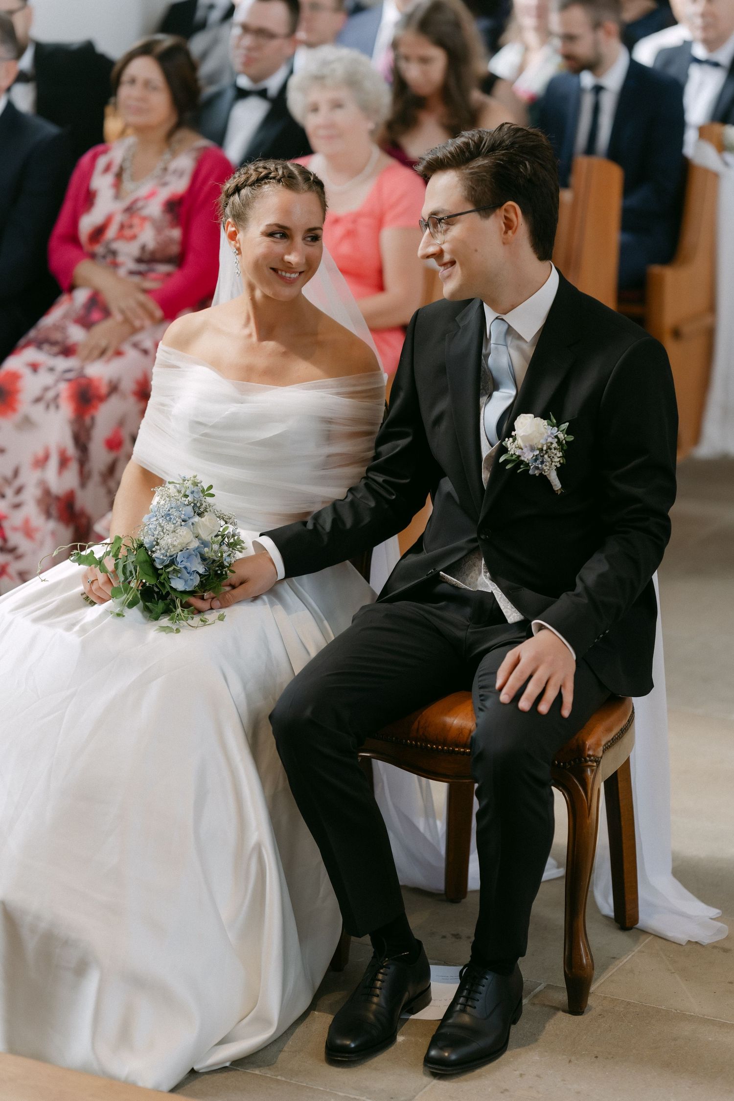Smiling bride in an off-shoulder gown holding a bouquet sits beside a groom in a dark suit during a church wedding ceremony with guests.