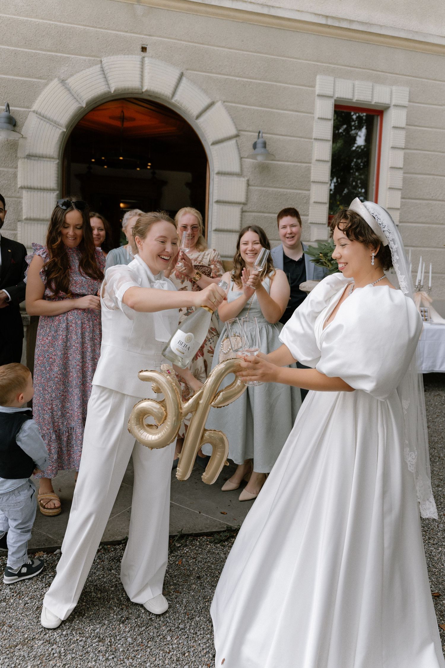 Two brides in white celebrate with champagne and gold balloons outside a venue surrounded by wedding guests.