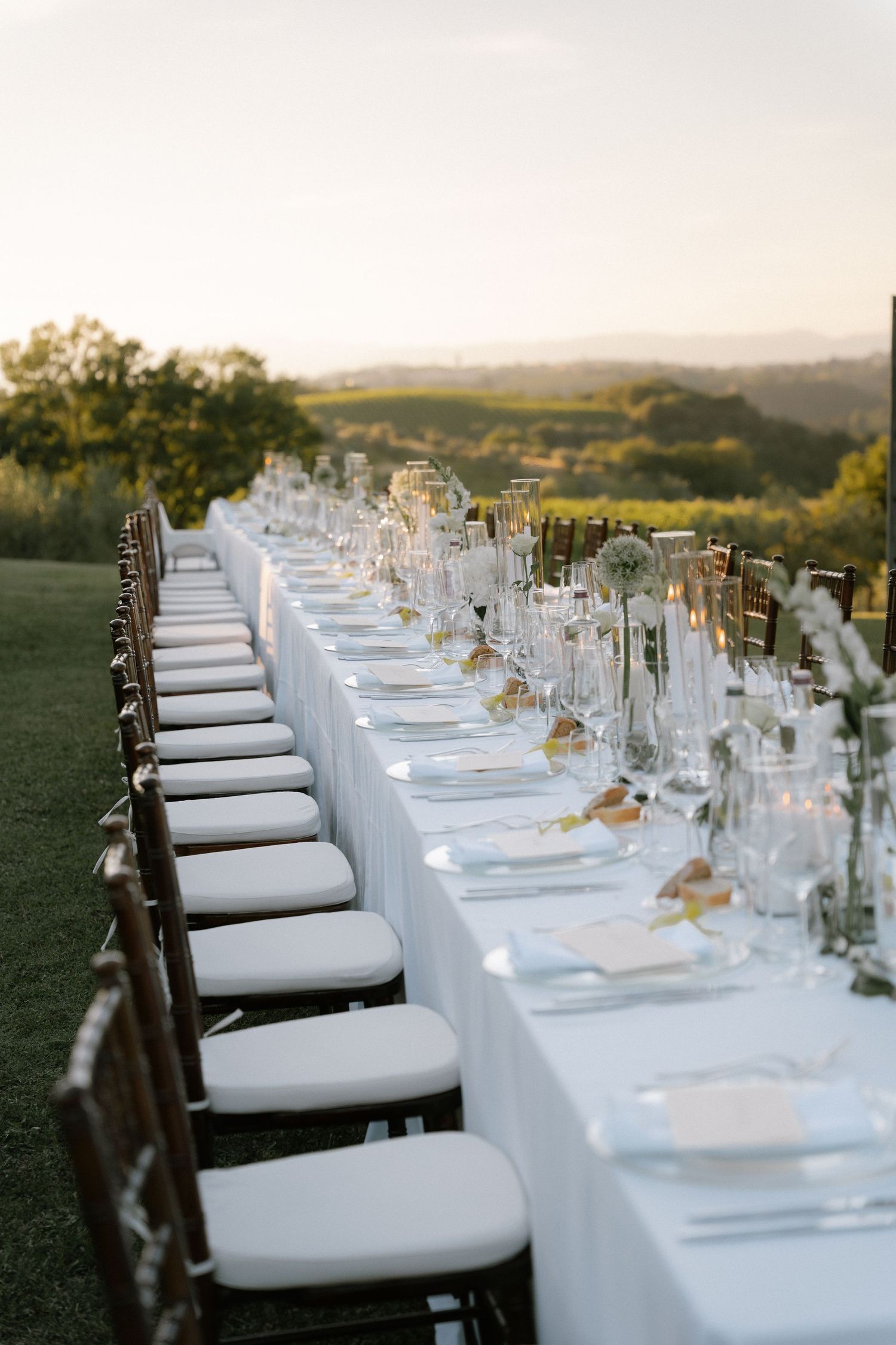 Elegant outdoor wedding reception with long white-draped table, chiavari chairs, floral centerpieces, and golden sunset views.