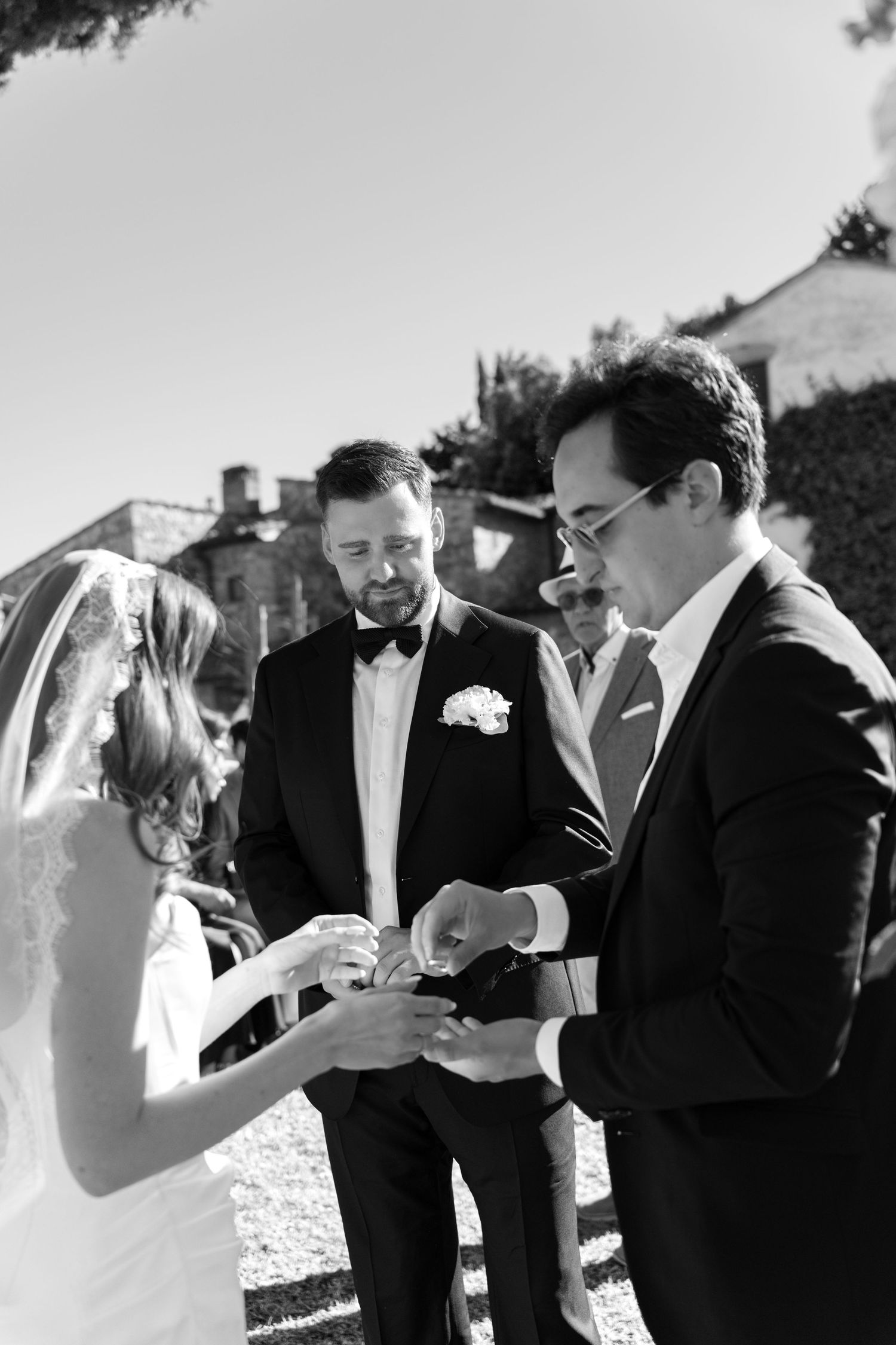 Black and white photo of groom placing wedding ring on bride's finger during outdoor ceremony.