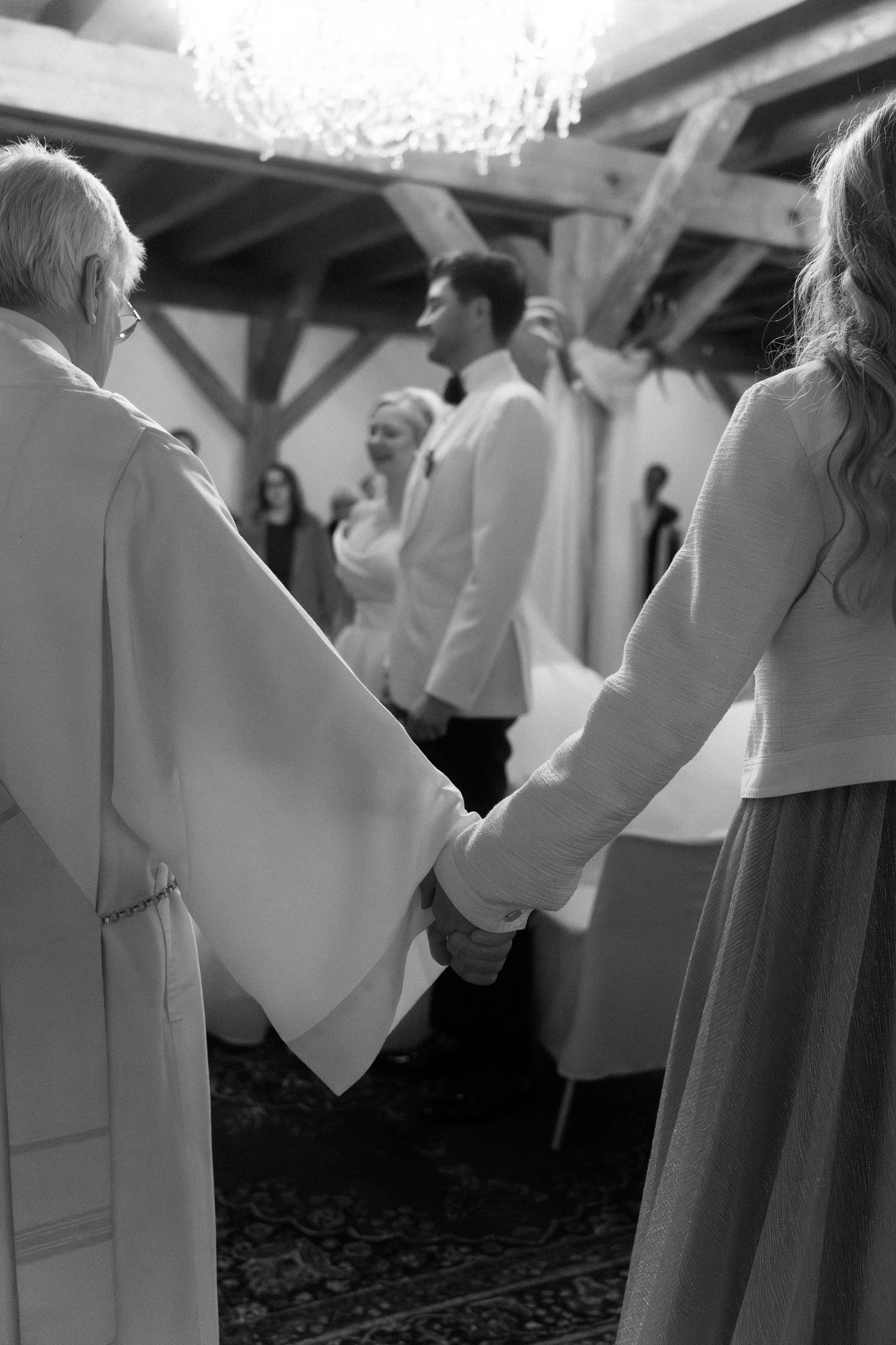 Black and white photo of people holding hands during a wedding ceremony in an indoor venue with exposed beams.
