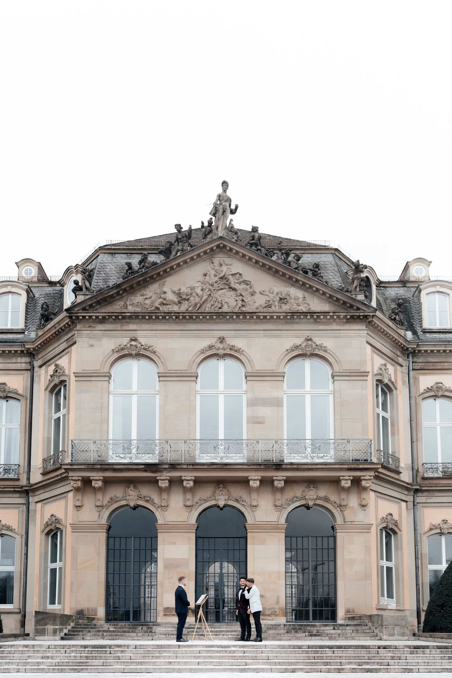 Ornate classical European building facade with triangular pediment and arched windows where people stand on grand steps.