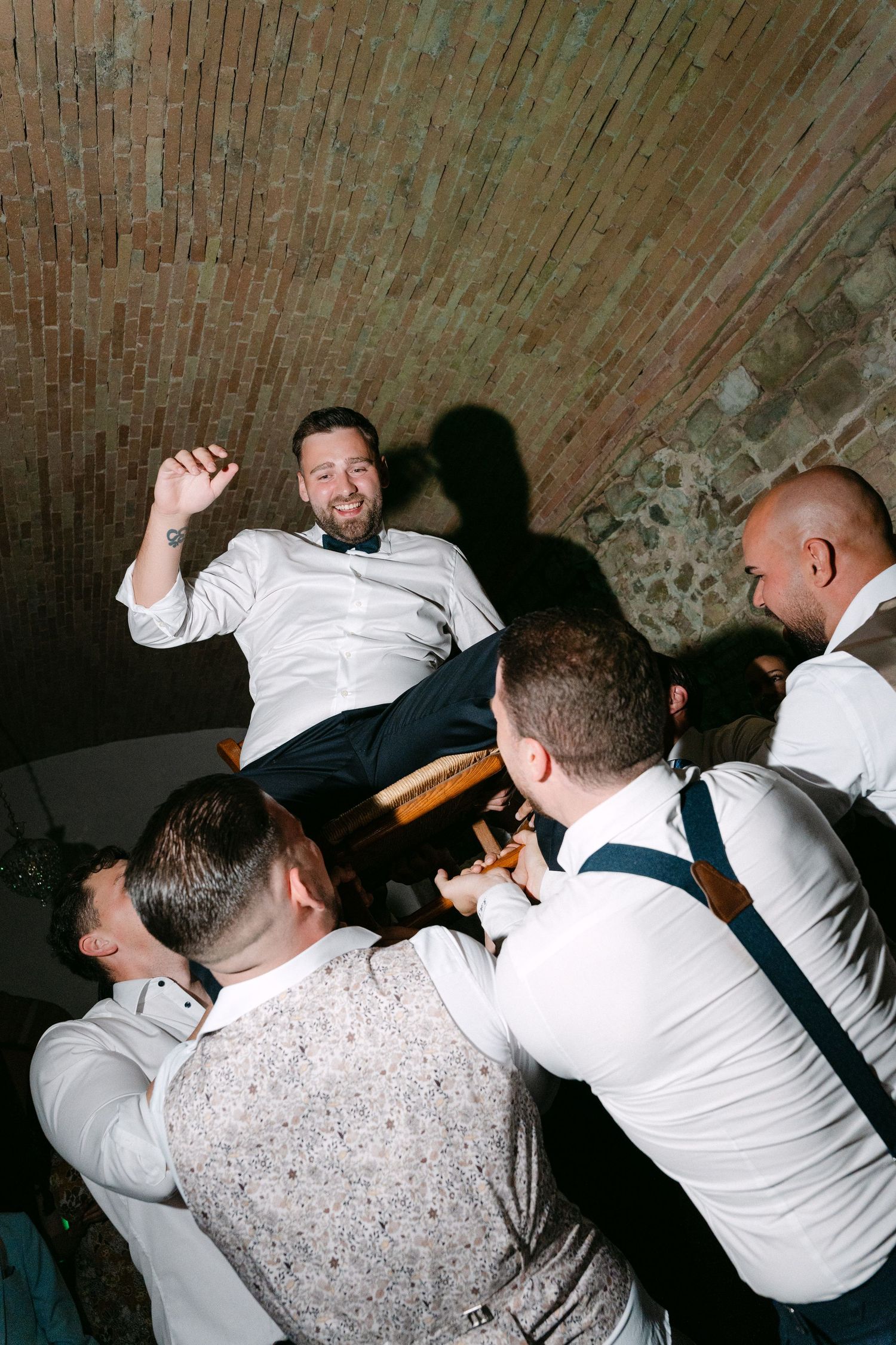 Groom being lifted by groomsmen during wedding reception celebration in rustic brick venue.
