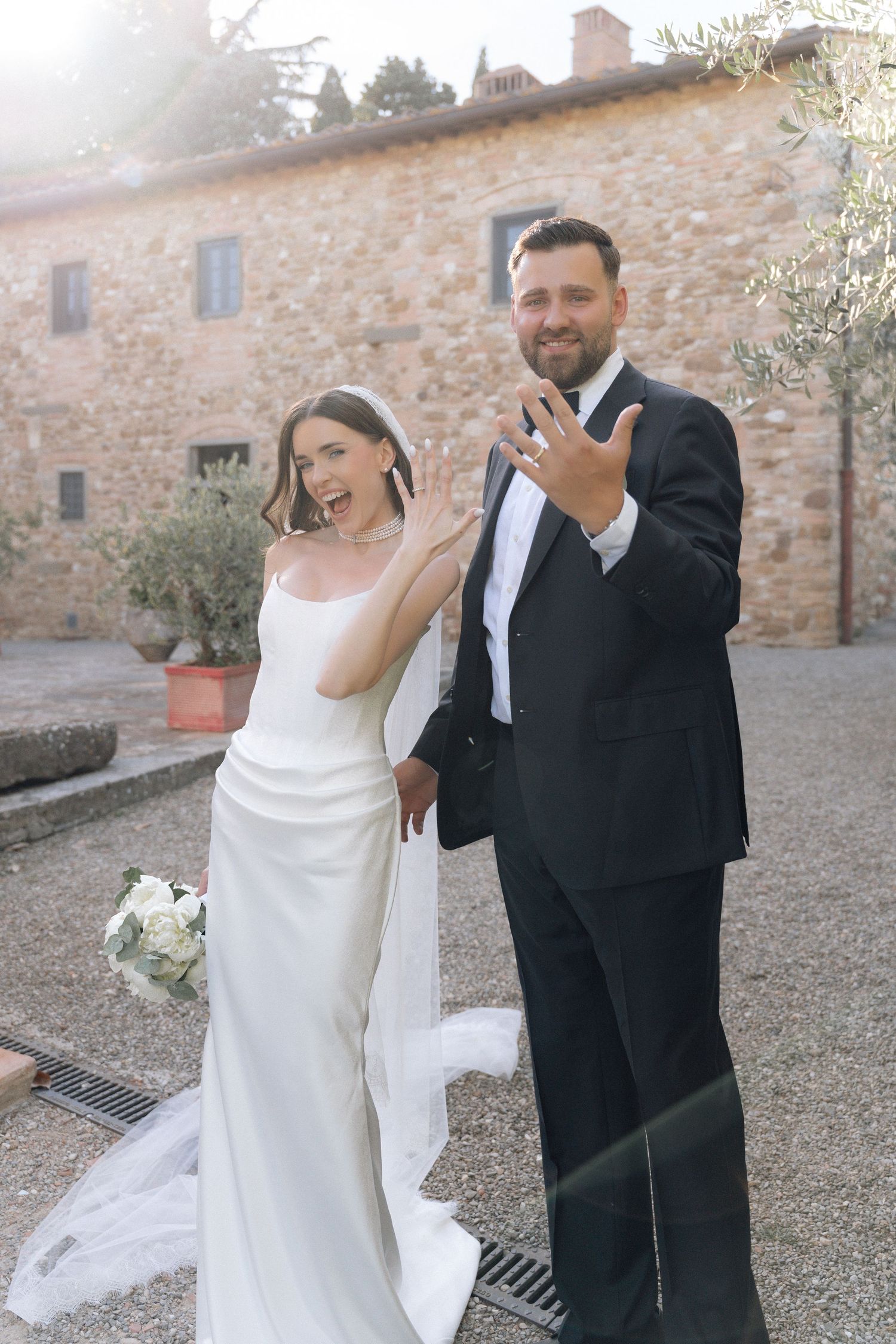 Newlywed couple in wedding attire showing off rings outside a Tuscan villa, bride in white gown holding bouquet.