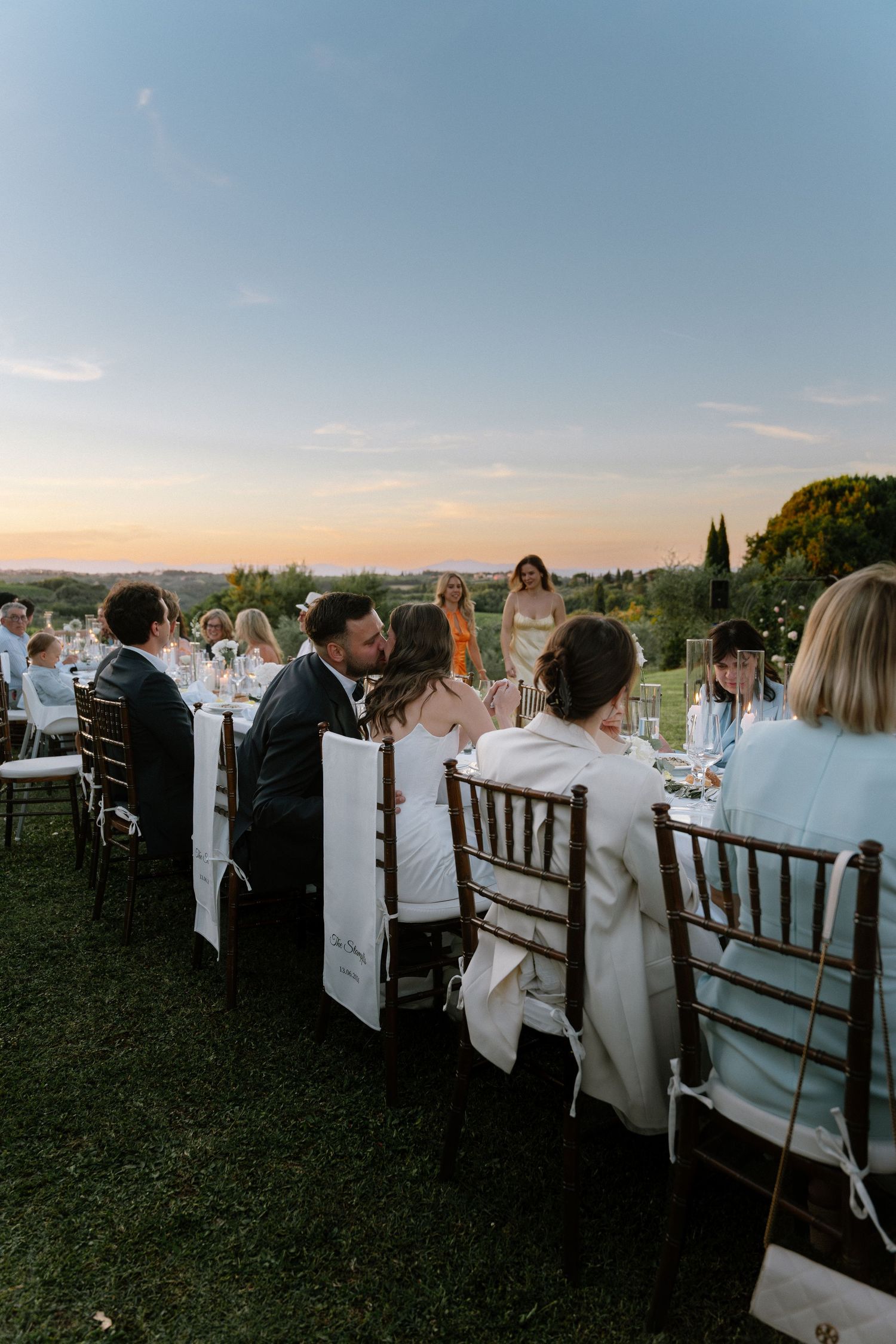 Couple sharing a kiss at an outdoor wedding reception dinner table during golden hour sunset.
