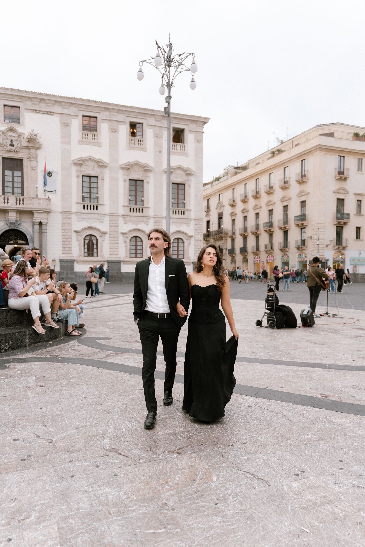 Elegant couple in black formal attire walking hand-in-hand through a grand European plaza with historic baroque buildings.