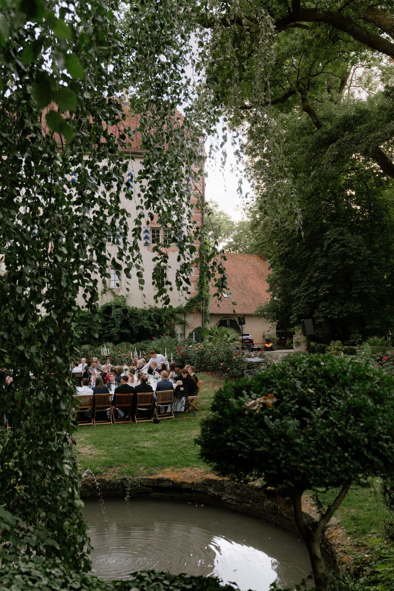 Wedding guests seated at a long outdoor table on a green lawn beside a small pond, framed by leafy trees and a historic manor house.