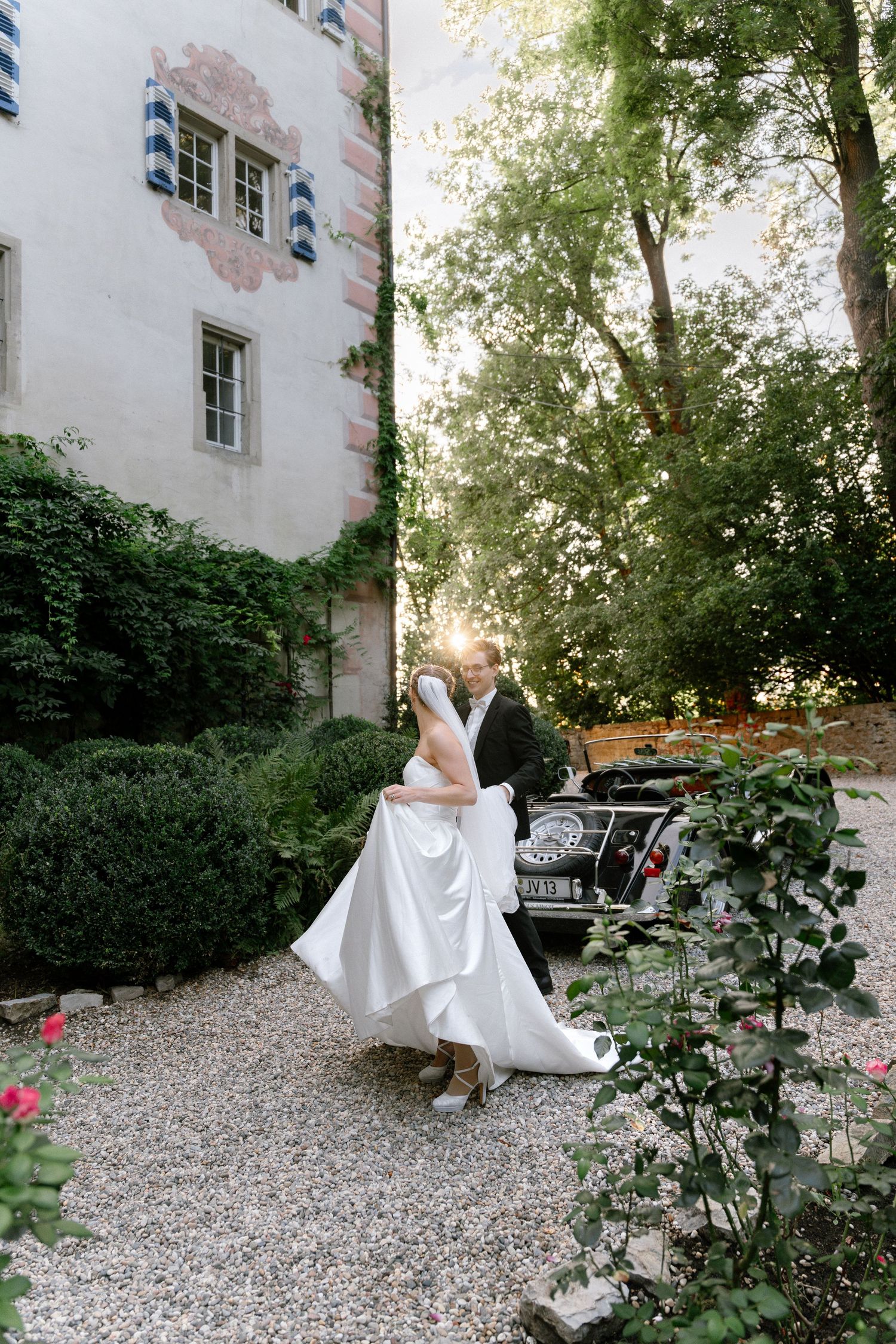 Bride in a flowing white gown embracing her groom on a gravel path near a vintage car and a castle-style building with striped shutters.
