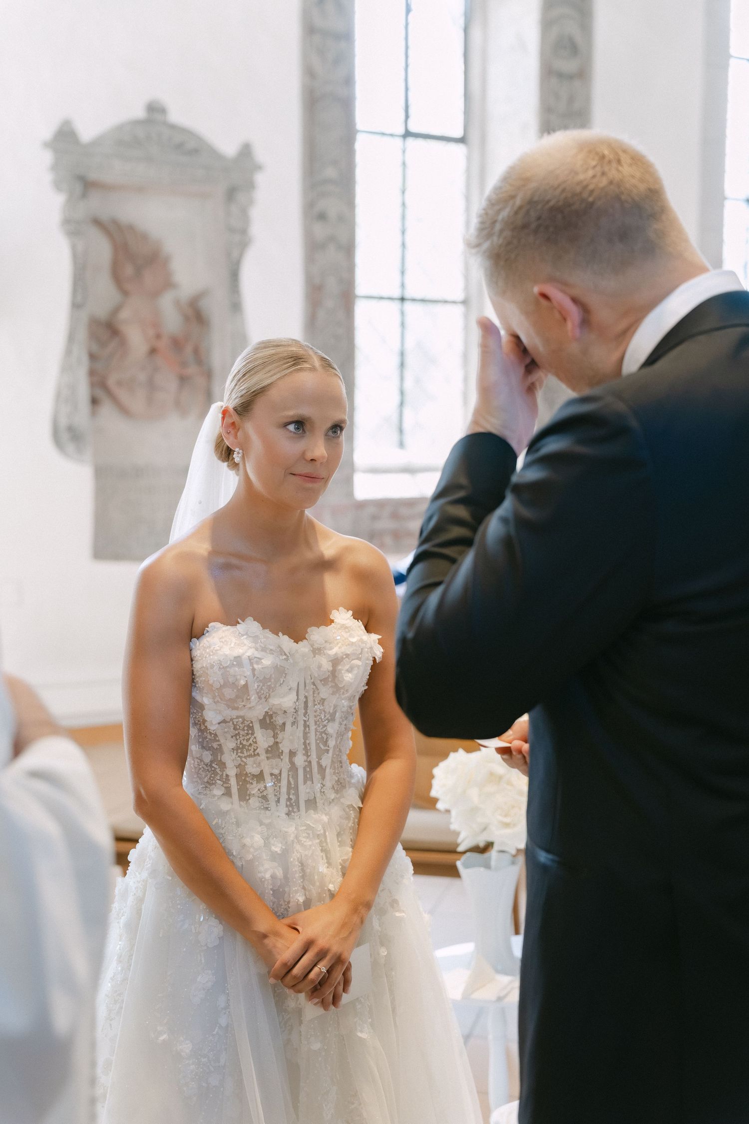 A bride in a strapless lace wedding gown and veil stands calmly as the groom wipes his eyes during a ceremony in a historic church.