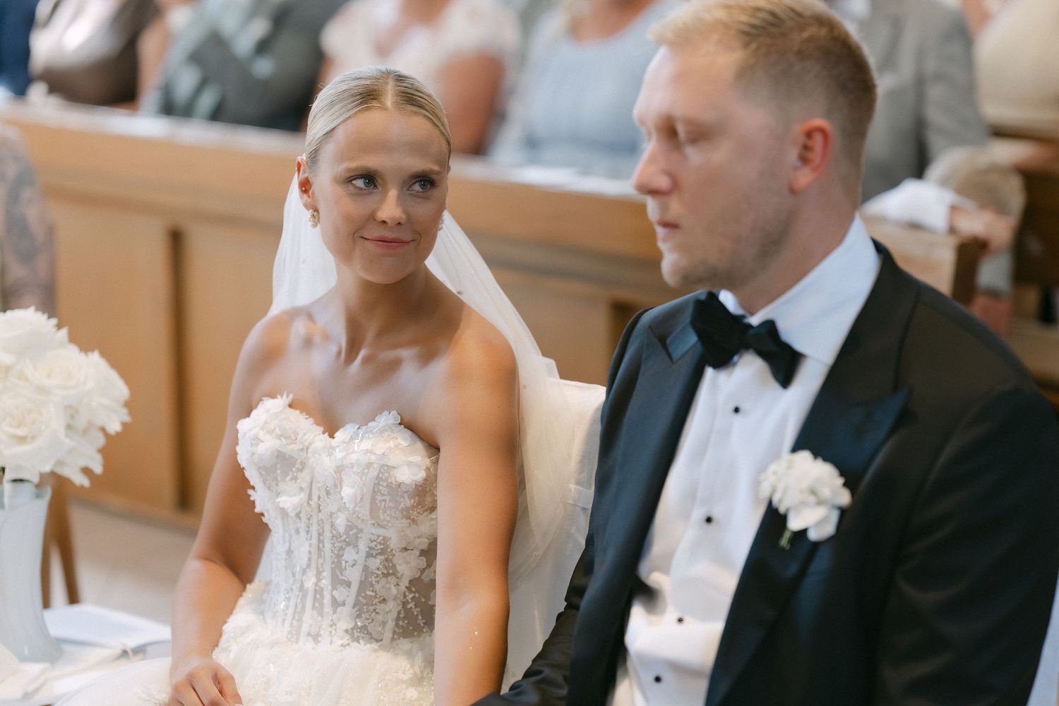 Bride in a white strapless lace wedding gown and veil sits beside a groom in a black tuxedo with bow tie during a wedding ceremony.