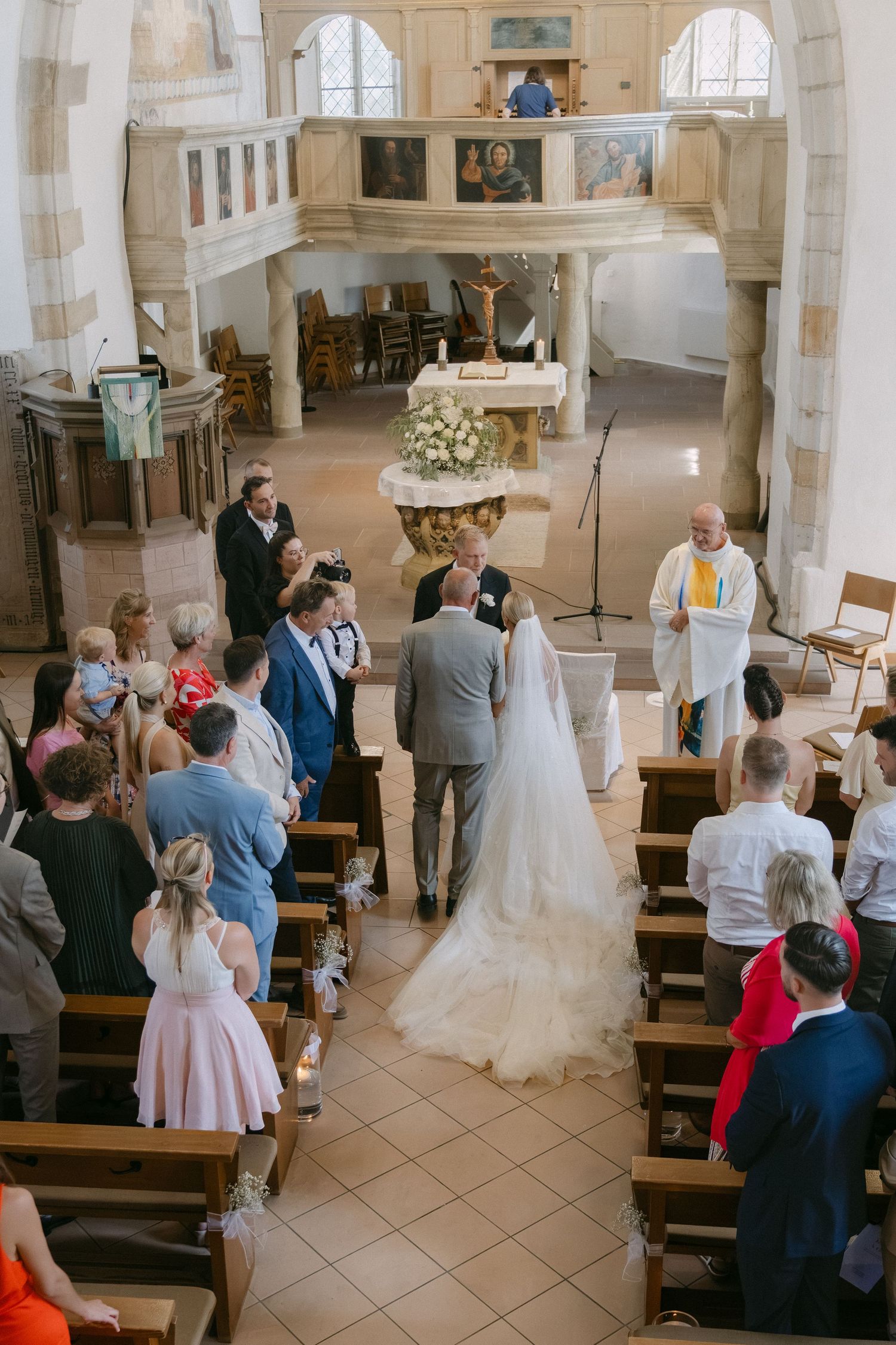 Bride in a long lace gown and groom stand before an officiant at the altar as wedding guests watch from wooden pews in a stone church.