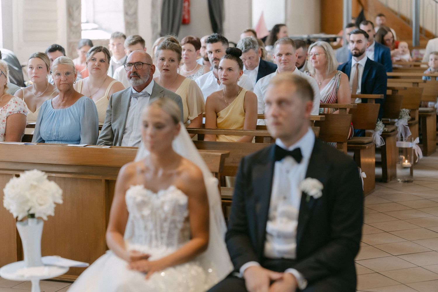 Bride in a white lace gown and groom in a black suit with bow tie sit in front of smiling wedding guests during a church ceremony.