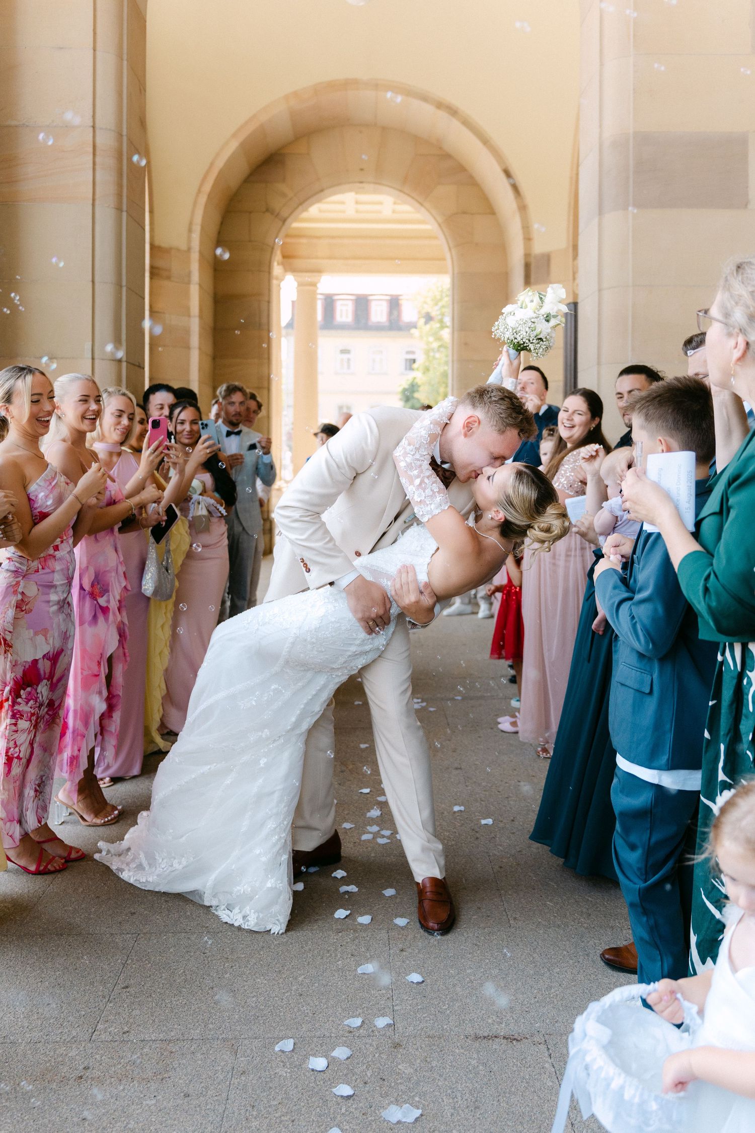 Wedding party guests toss confetti and celebrate under an outdoor colonnade during a festive reception exit.