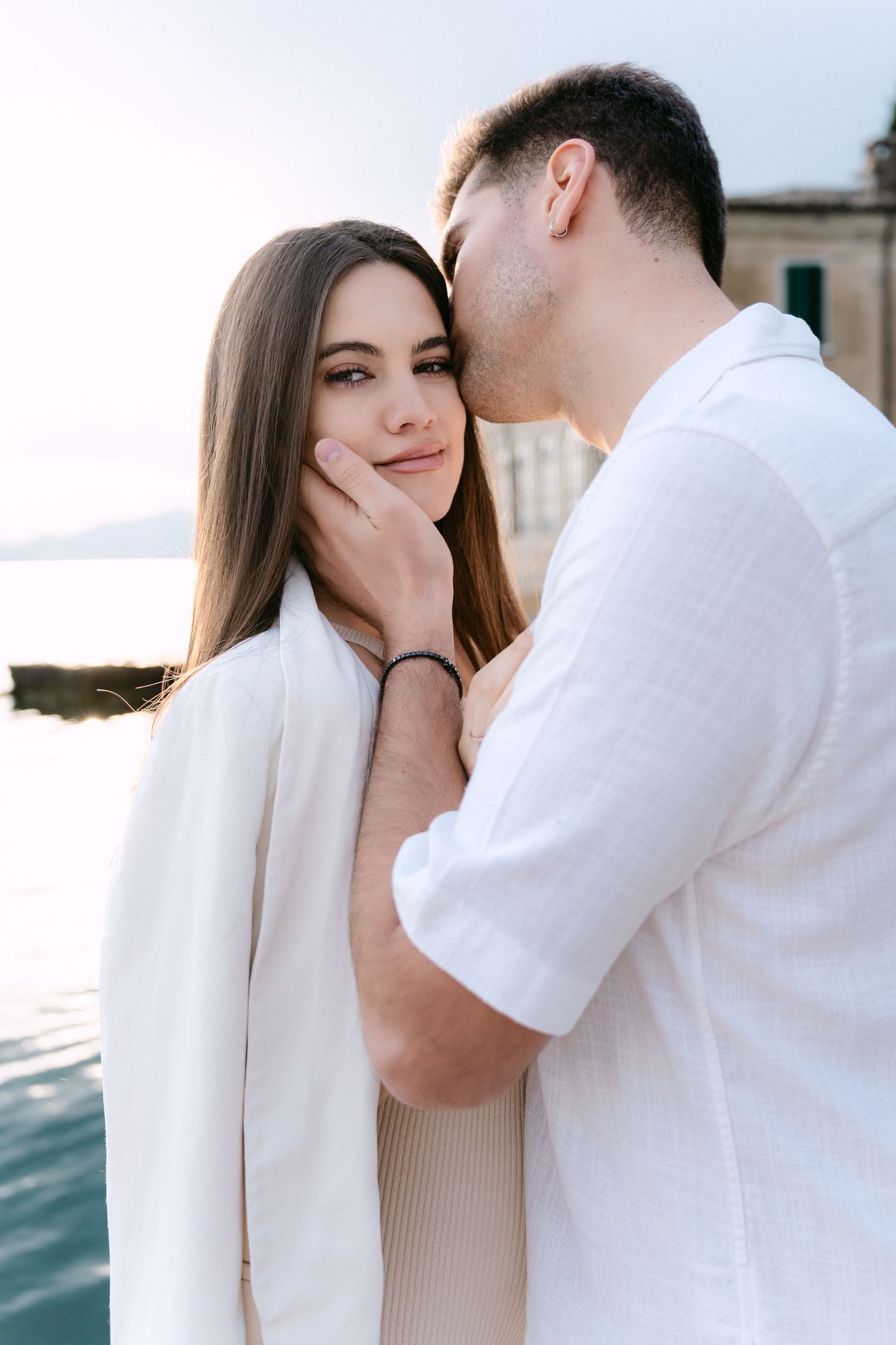 A romantic couple in white outfits share tender moments together in a series of intimate photos by the water.