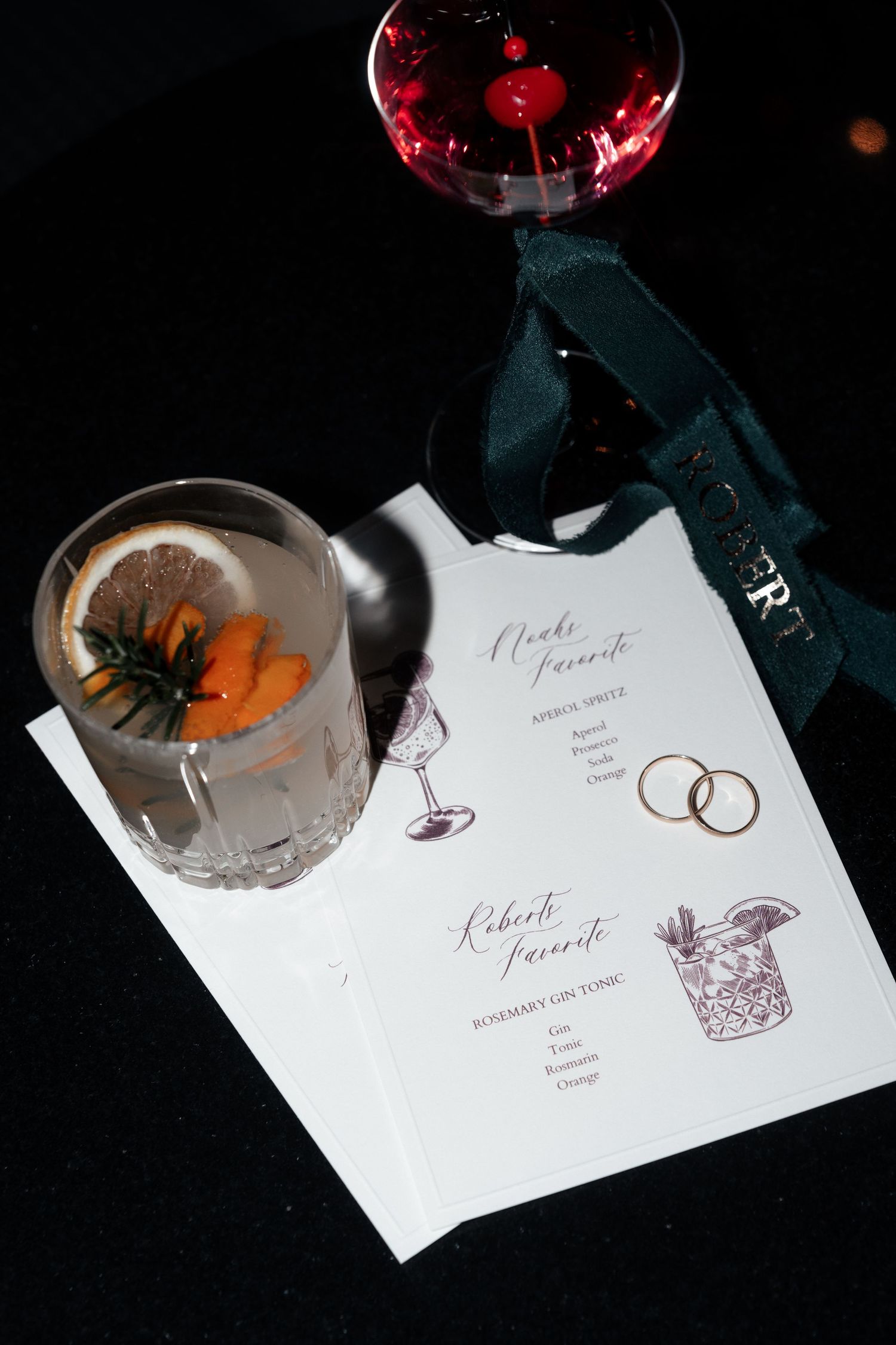 Cocktail menu with wedding rings and two drinks garnished with herbs on a dark surface.