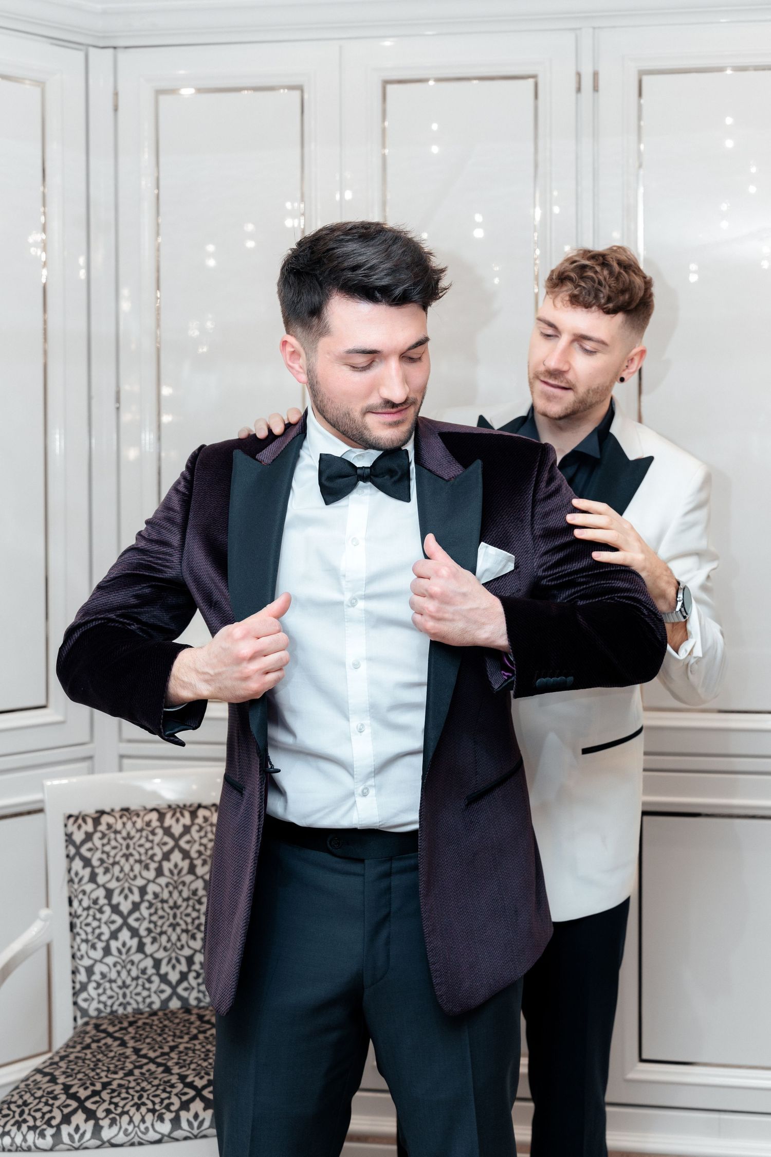 Two men in formal attire getting ready, one in a black tuxedo adjusting his bow tie while another assists.